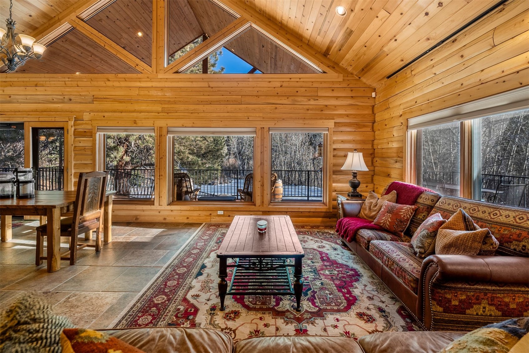 Log-cabin living room with vaulted wood ceiling, patterned sofa, and windows overlooking snowy woods.