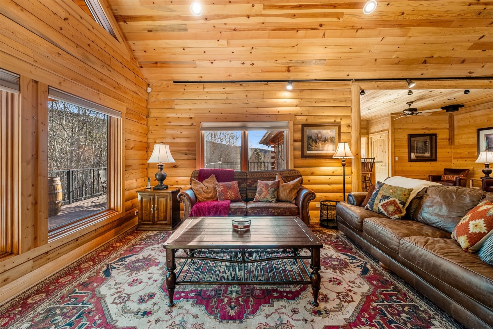 Log cabin living room with wood walls, leather sofas, a patterned rug, and windows facing a deck.