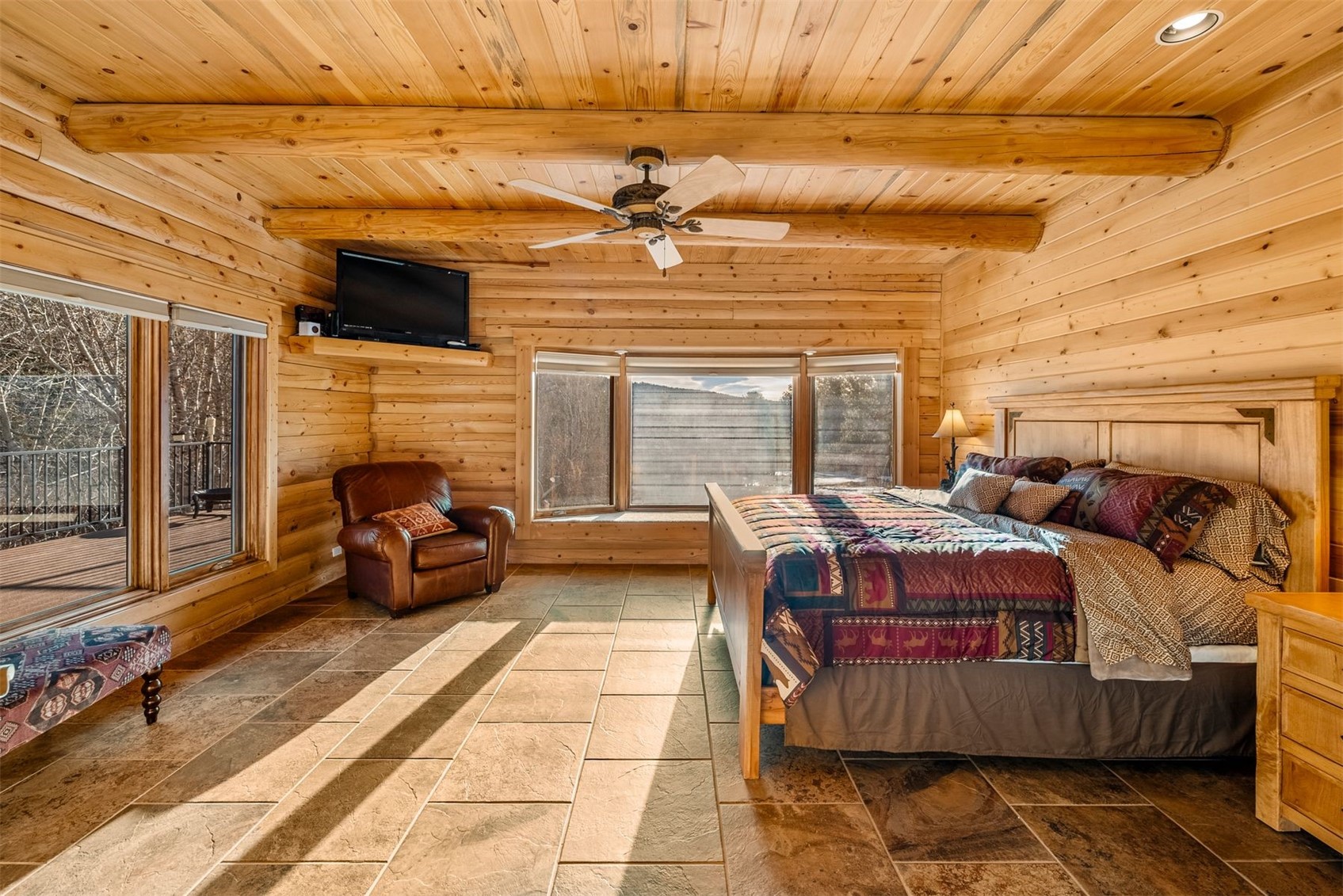 Log bedroom with exposed beams, a patterned bedspread, tiled floor, and windows overlooking a deck.