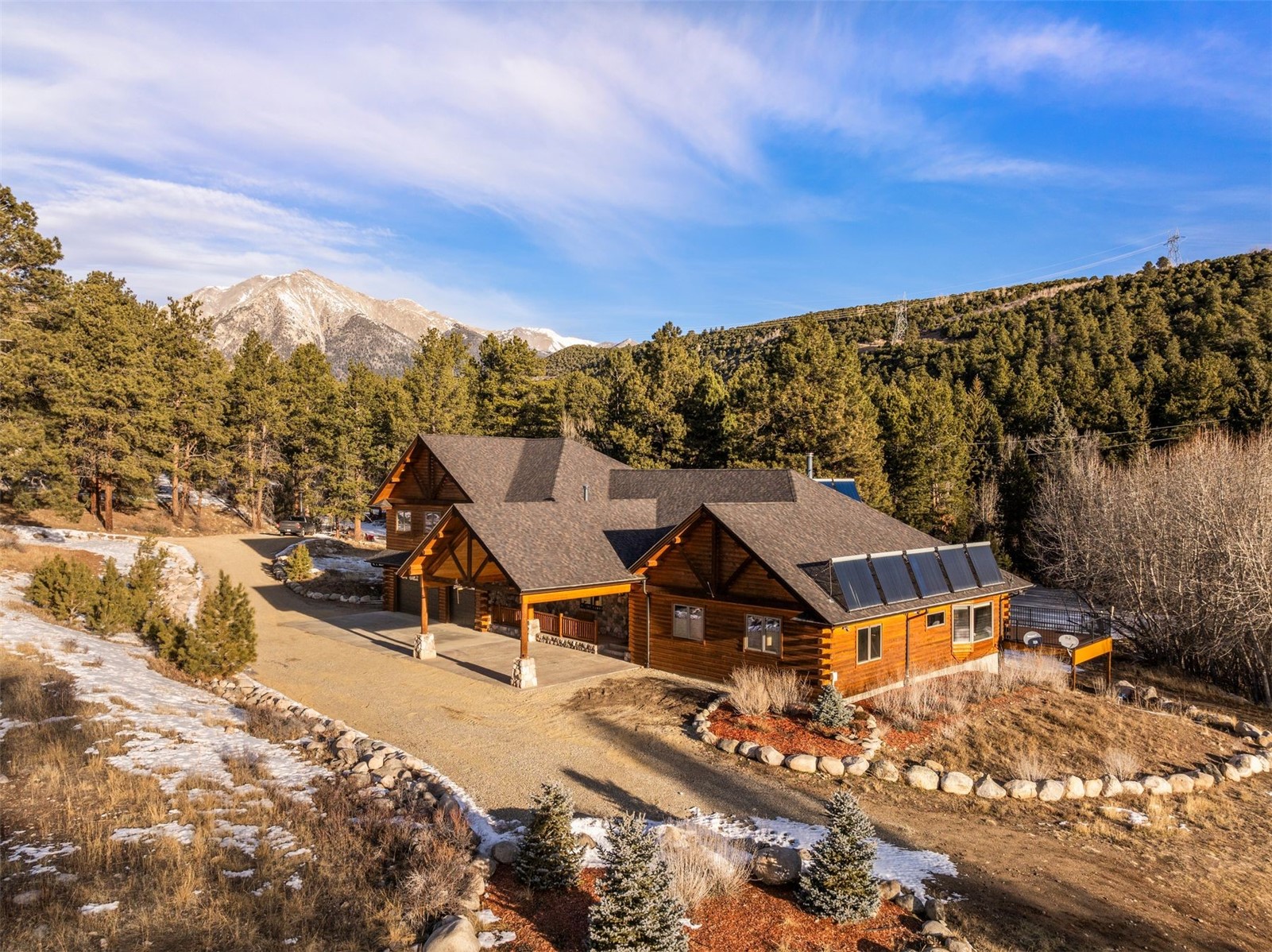 Log cabin with roof-mounted solar panels, a curved driveway, and snowy mountain views.