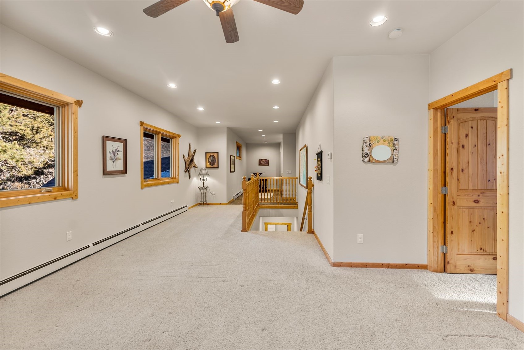 Carpeted upstairs landing with a wood stair railing, ceiling fan, and windows overlooking trees.