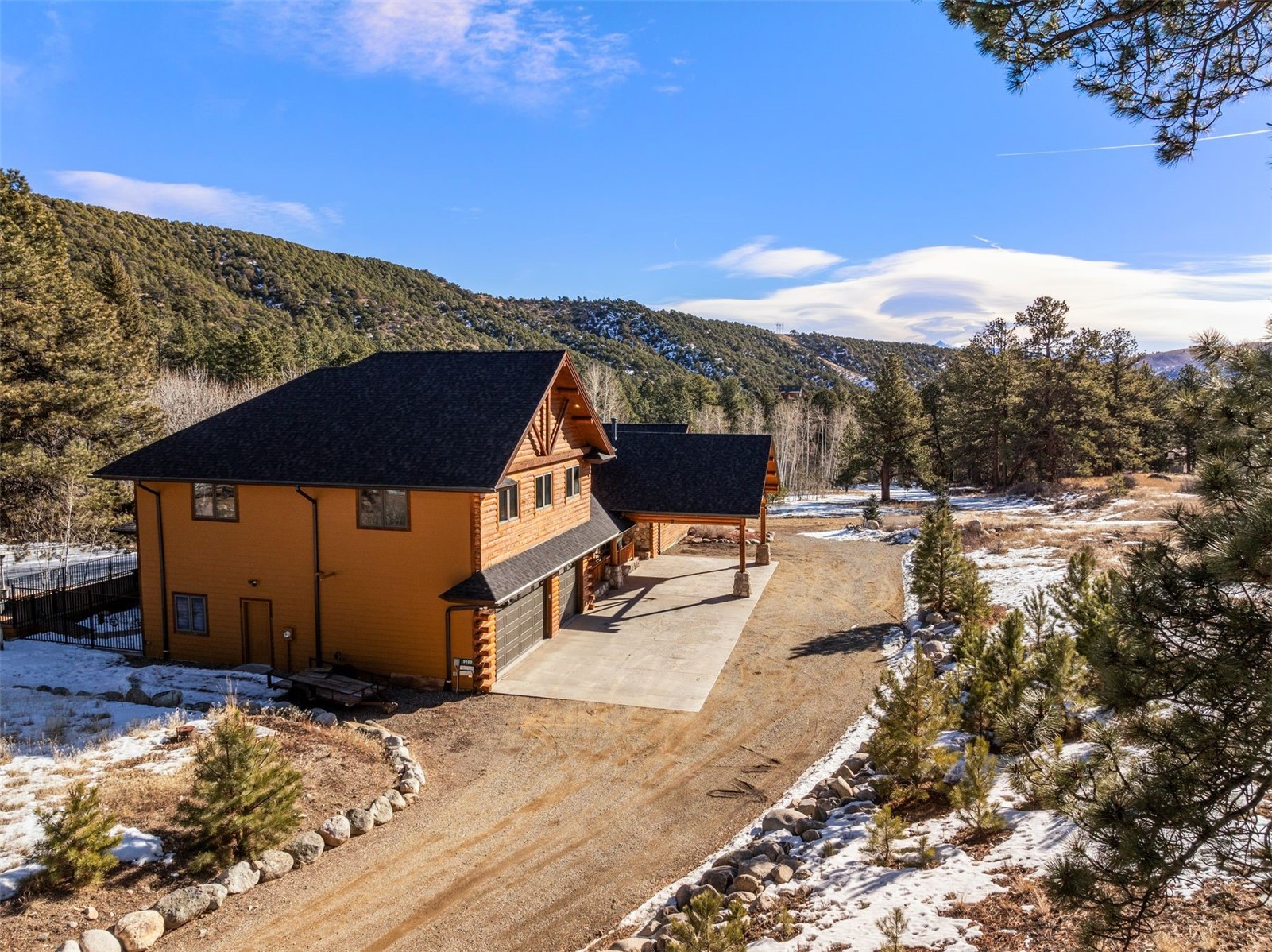 Log-sided mountain home with an attached garage, broad driveway, wooded hills, and patches of snow.