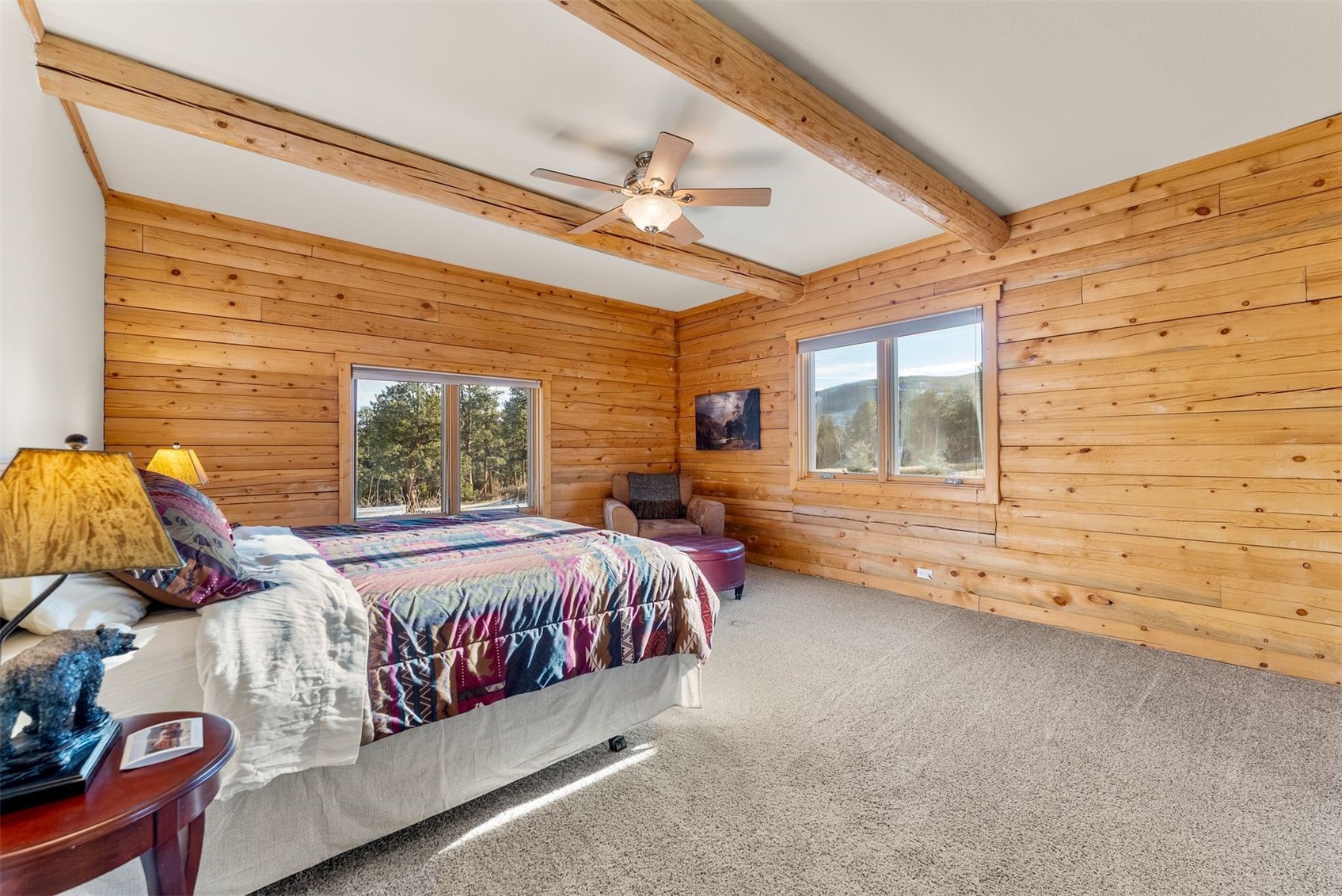 Wood-paneled bedroom with exposed beams, a colorful bedspread, and large windows overlooking trees.