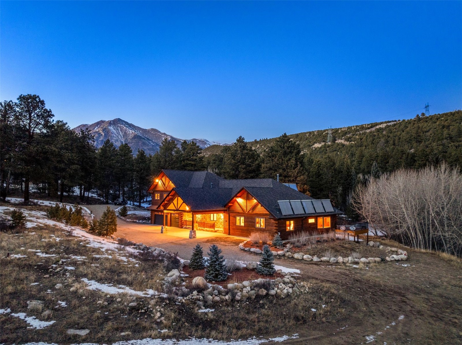 Log-and-stone mountain home with solar panels, glowing warmly at dusk beneath snow-dusted peaks.
