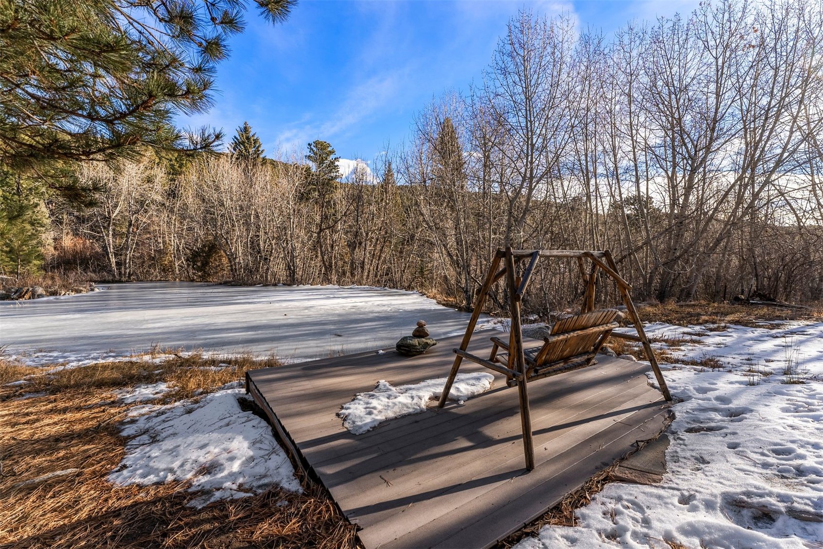 Wooden deck swing beside a frozen pond, surrounded by snowy ground and leafless trees.