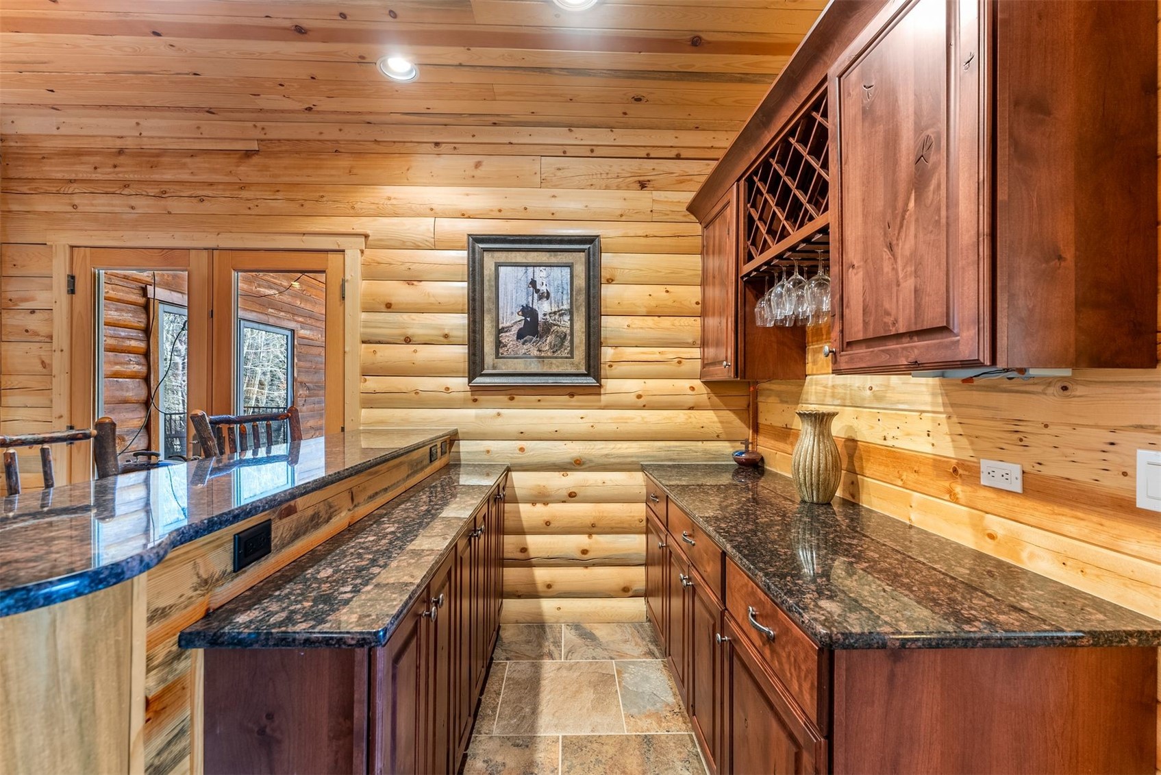 Log-home wet bar with granite counters, warm wood cabinetry, and a wine-glass rack.