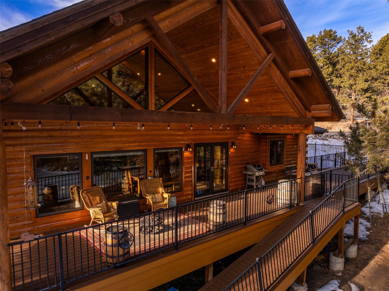 Log cabin with a tall timber gable and furnished raised deck amid patches of snow.