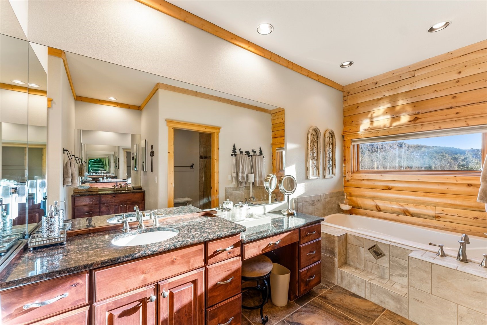Bathroom with double sinks, wood cabinetry, tiled bathtub, and a wide window overlooking wooded hills.