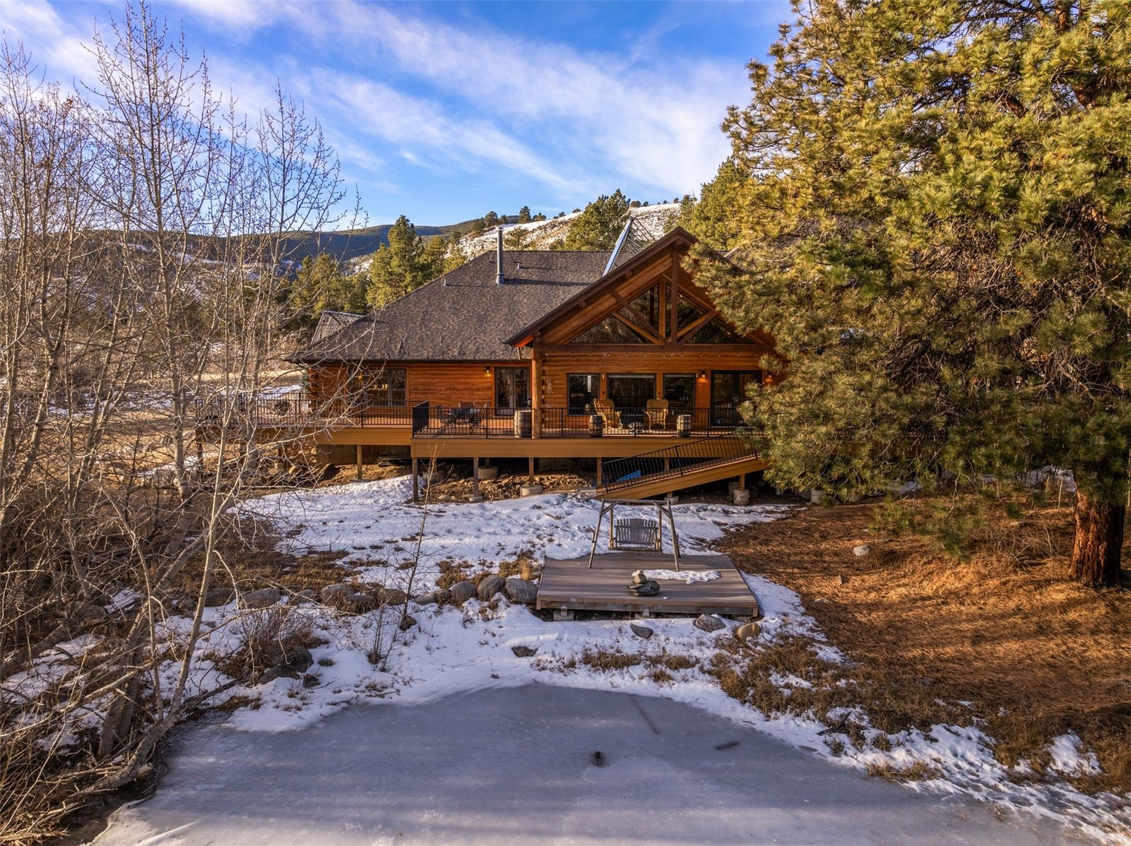 Wood mountain home with a timber-truss roof and raised deck beside a frozen pond.