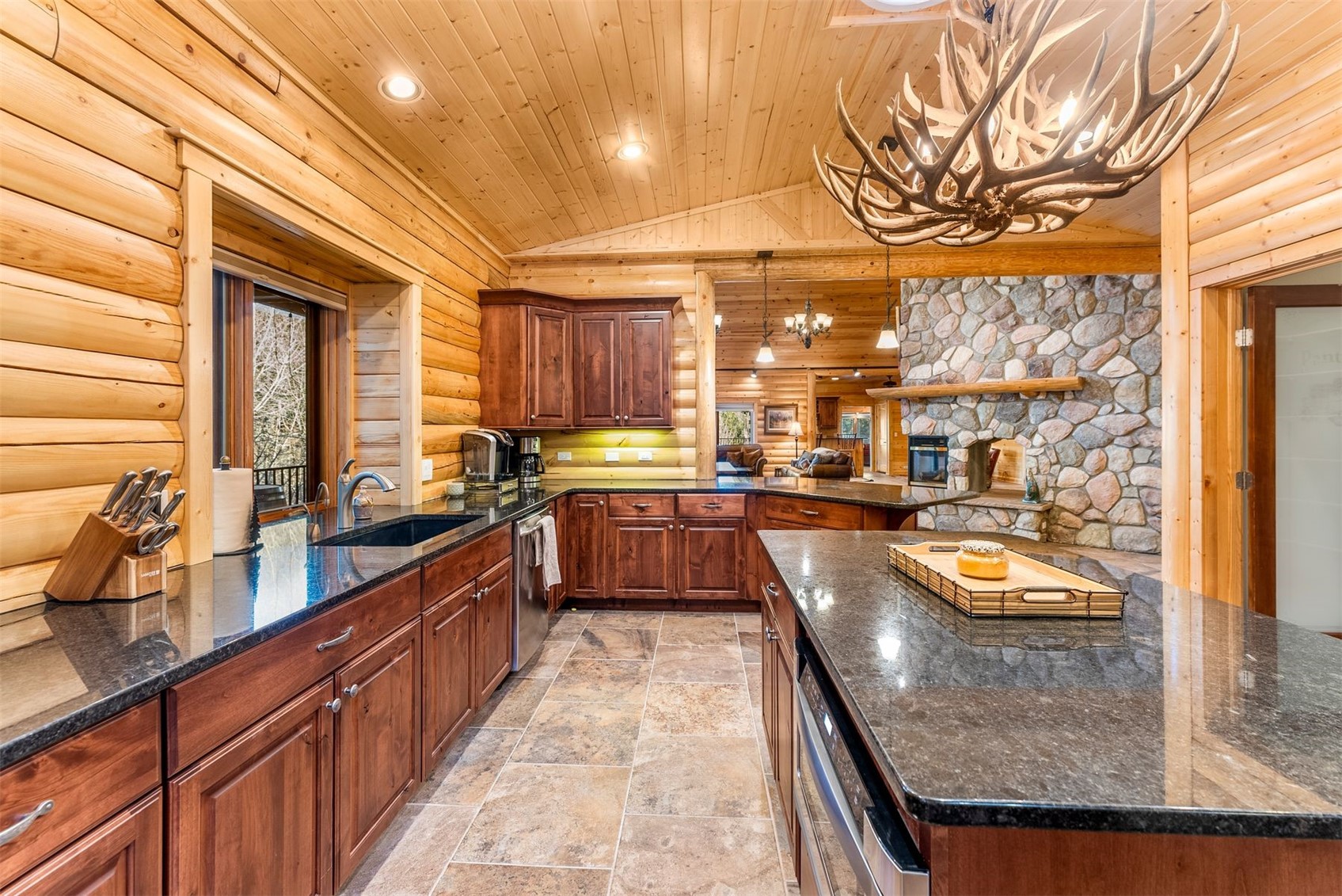 Log-home kitchen with dark wood cabinets, granite island, and antler chandelier.