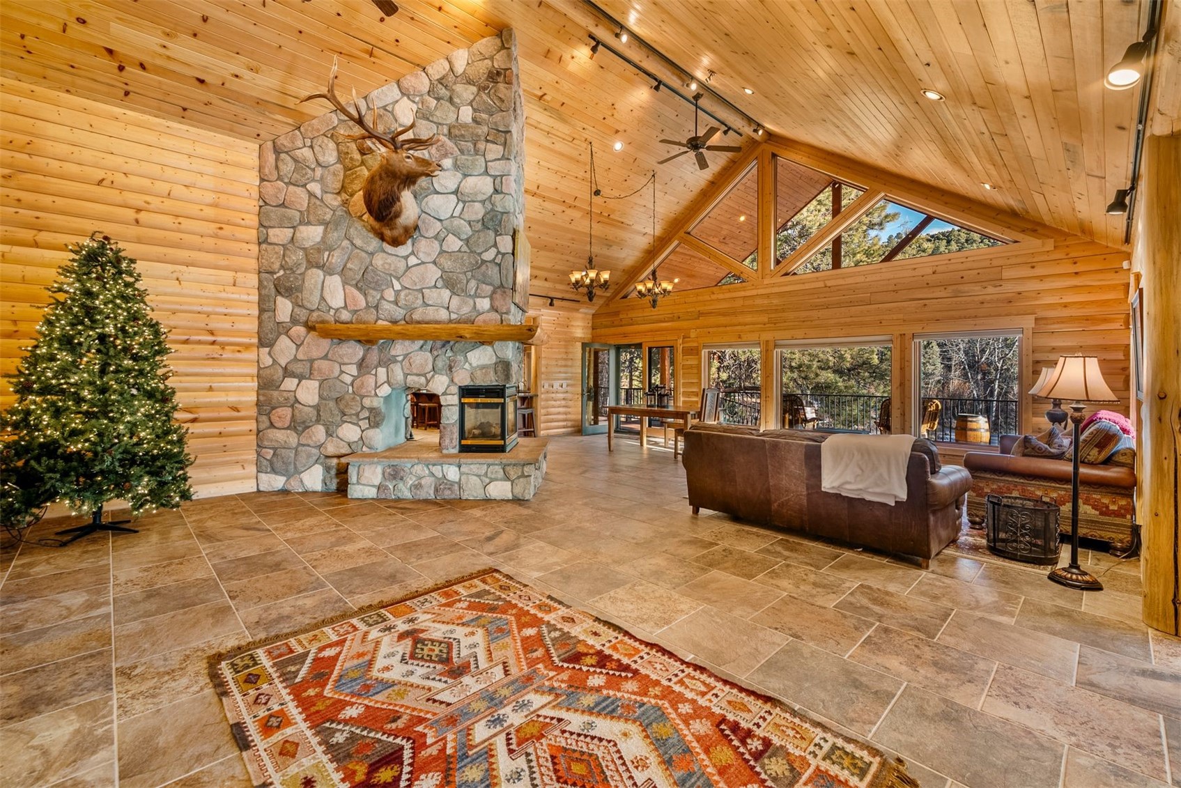 Vaulted pine great room with a stone fireplace, elk mount, and large windows overlooking trees.