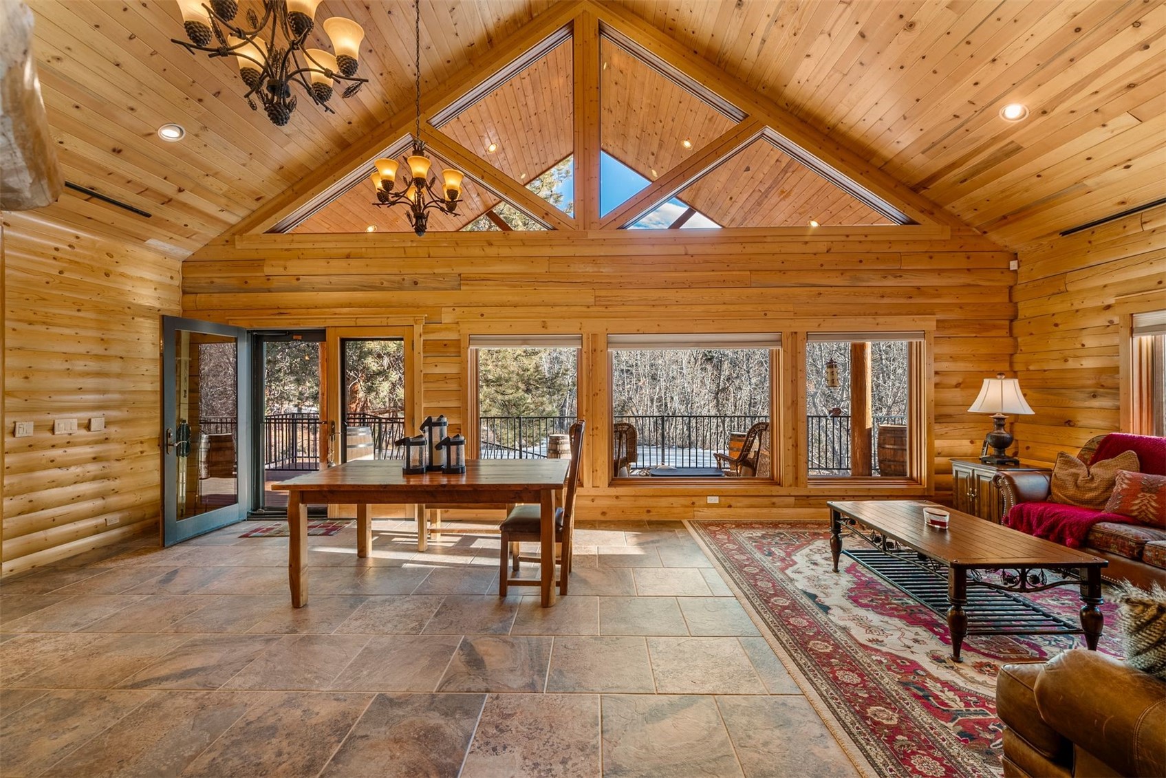 Furnished log-home great room with vaulted pine ceiling and triangular clerestory windows overlooking a wooded deck.