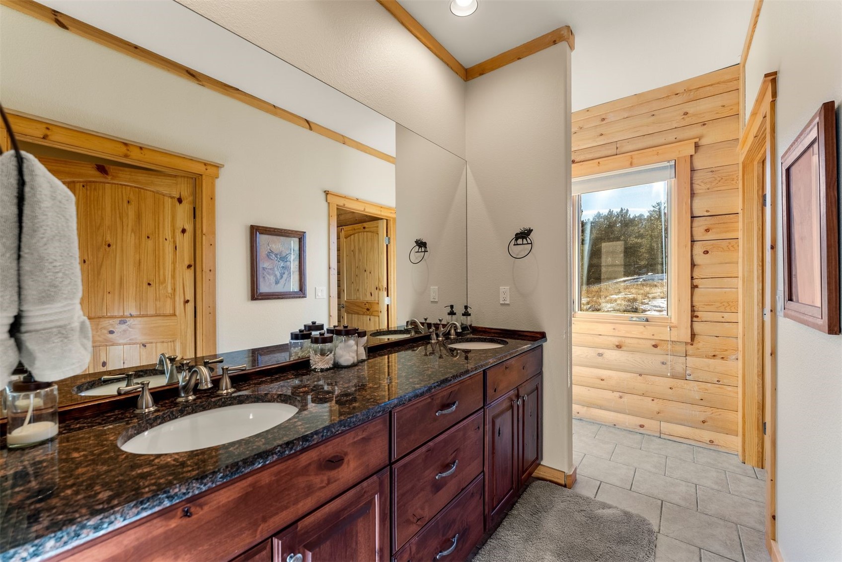 Bathroom vanity with two sinks, dark granite counter, knotty pine trim, and a window overlooking snowy ground.