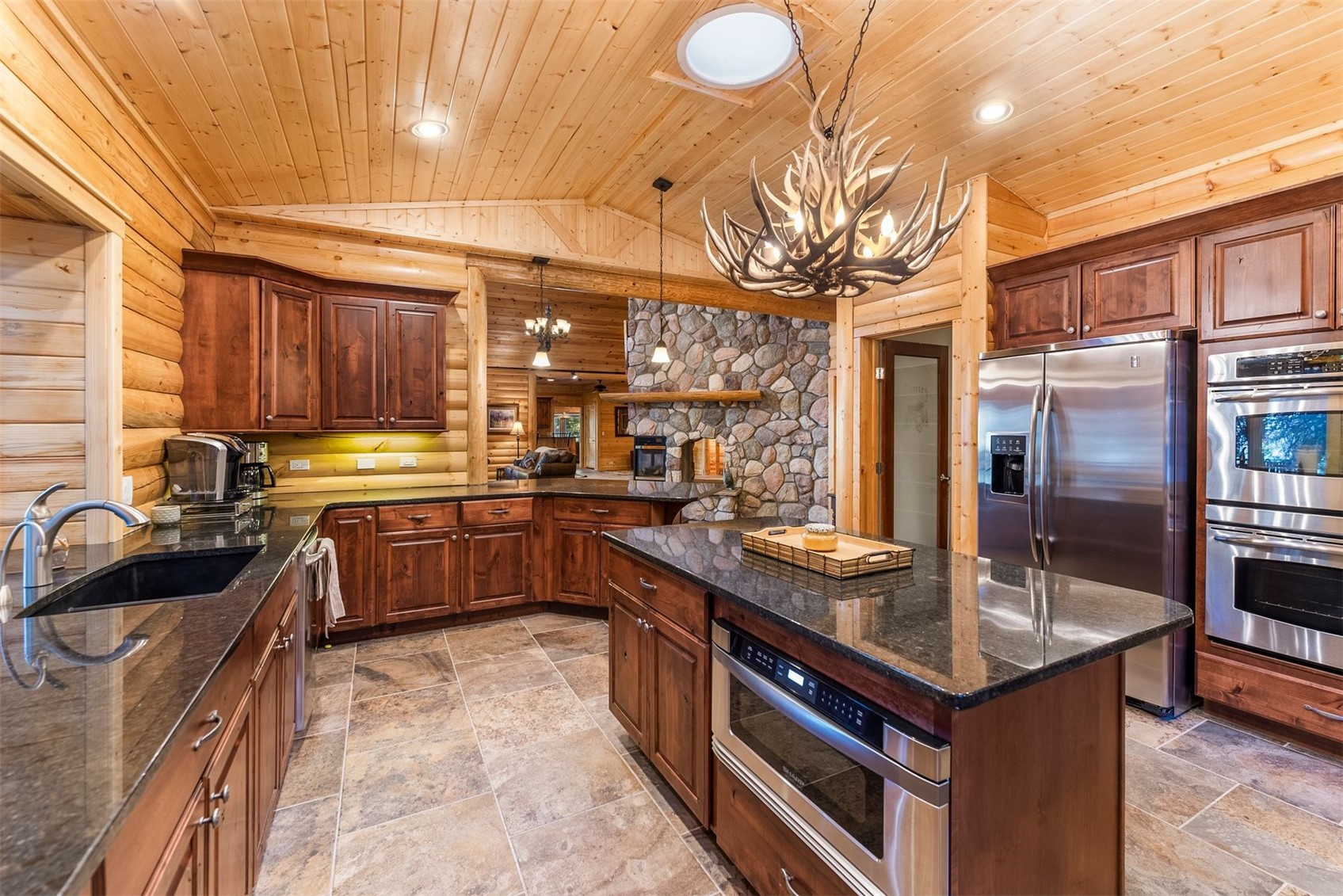 Log-cabin kitchen with granite island, stainless steel appliances, and antler chandelier.