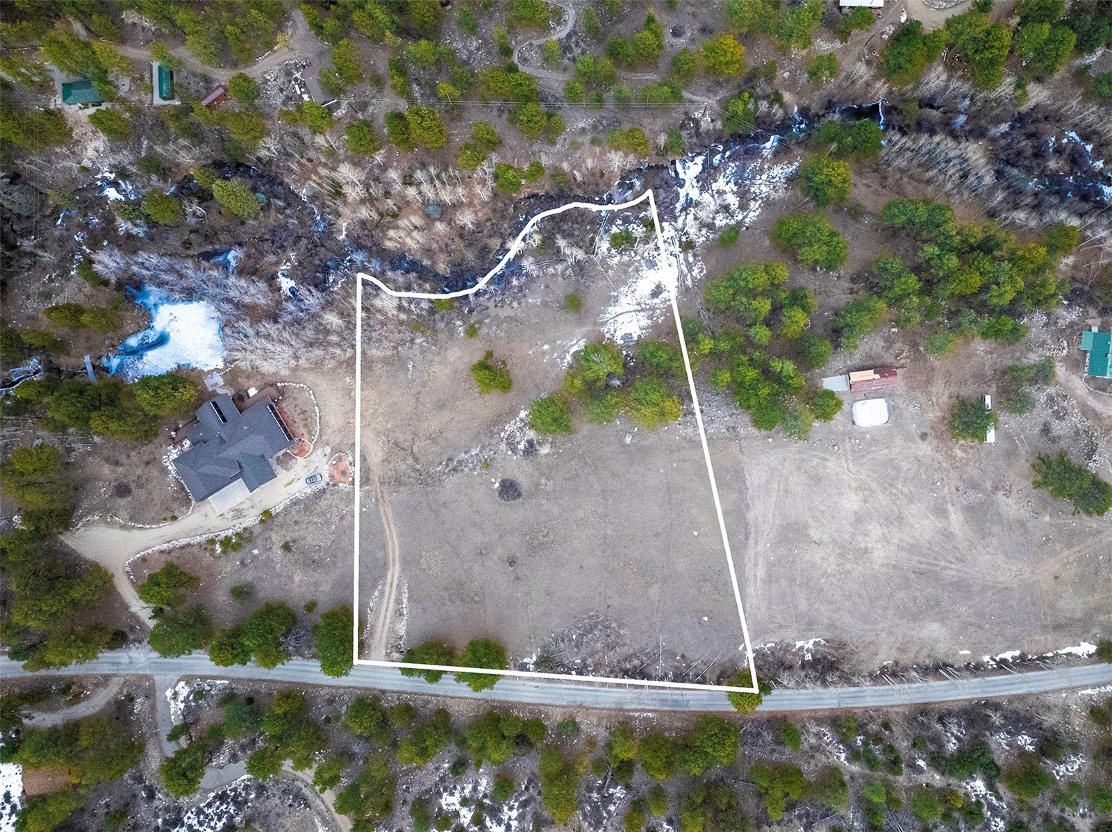 Aerial view of a rural parcel outlined in white beside a road, with scattered trees and patches of snow.
