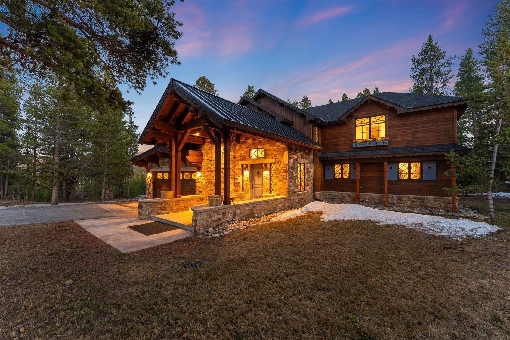 Rustic mountain home with stone entry, timber siding, glowing windows, and snow patches at dusk.
