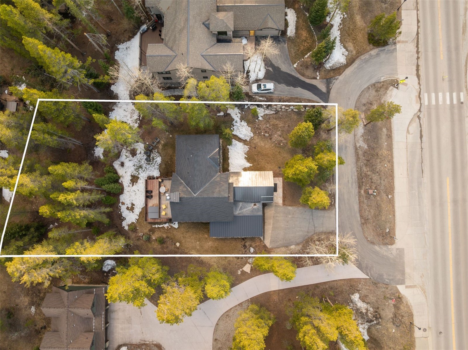 Aerial view of a house and wooded lot outlined in white, with a deck, driveway, and roadside access.