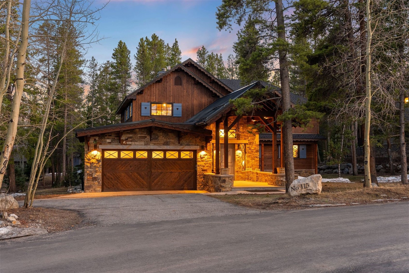 Mountain home with stone and wood exterior, warmly lit at dusk among trees.