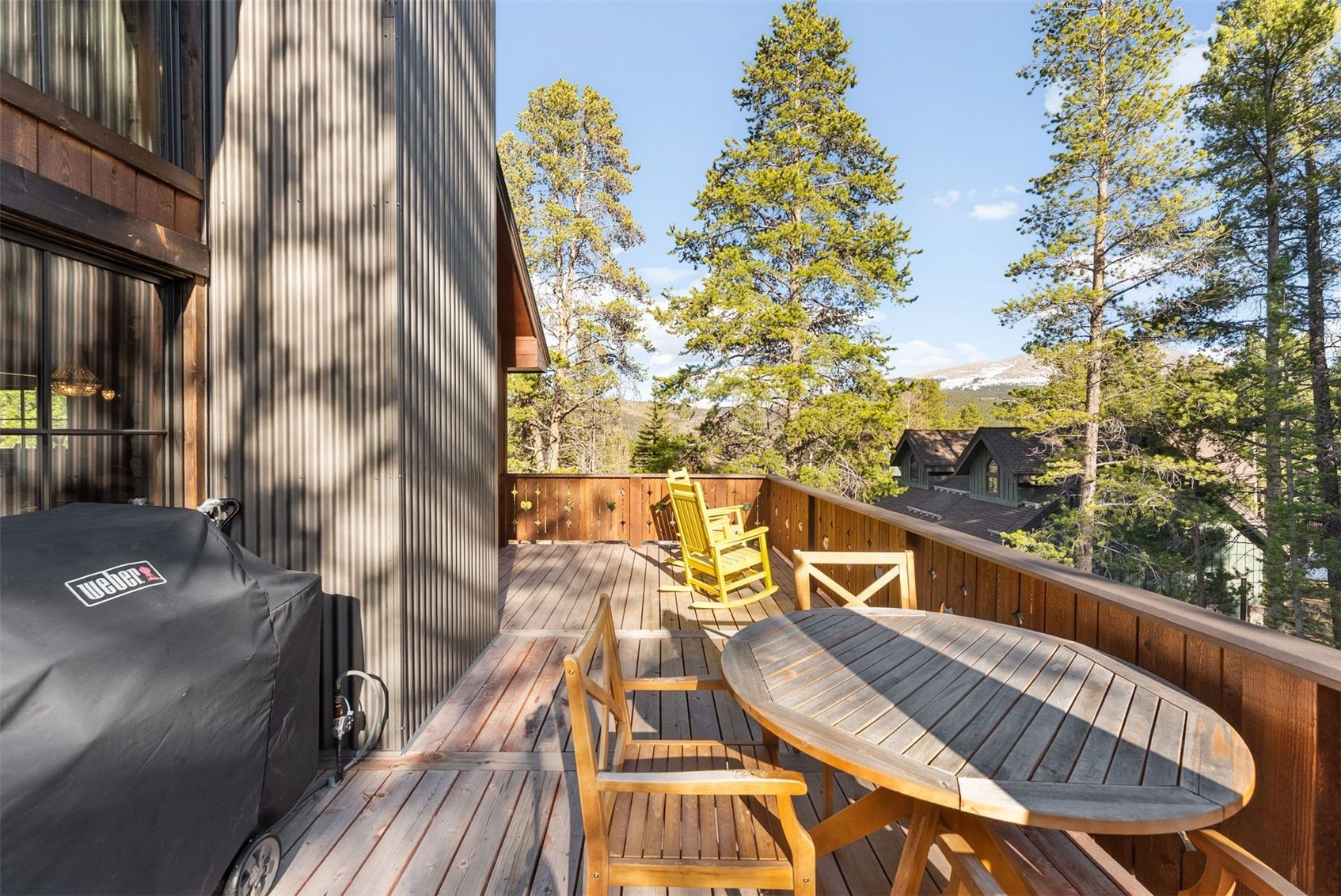 Wooden deck with outdoor dining set and grill overlooking pine trees and snow-dusted mountains.