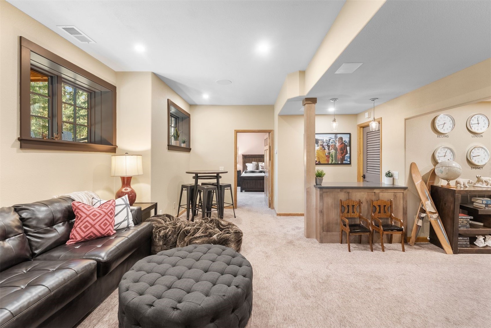 Lower-level living room with leather sectional, tufted ottoman, and a small bar beside world clocks.