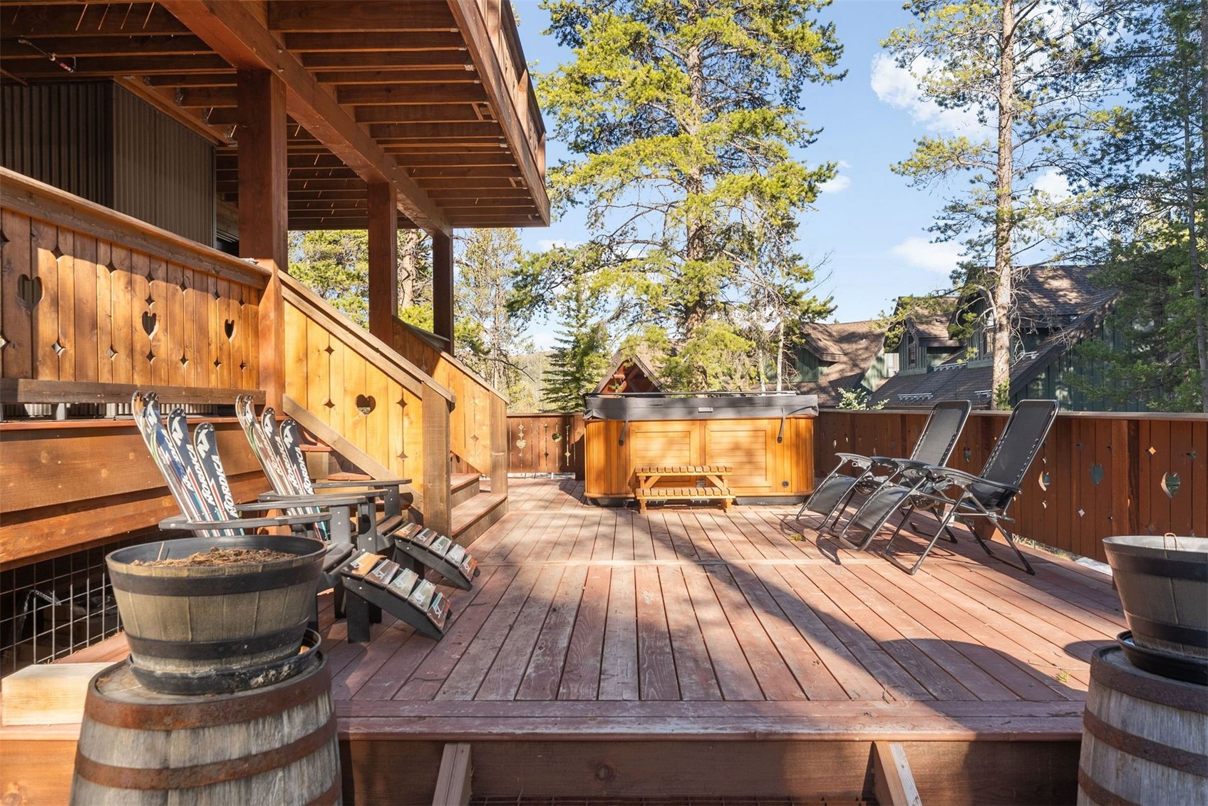 Wooden deck with a hot tub, lounge chairs, and heart-cutout railing among pine trees.