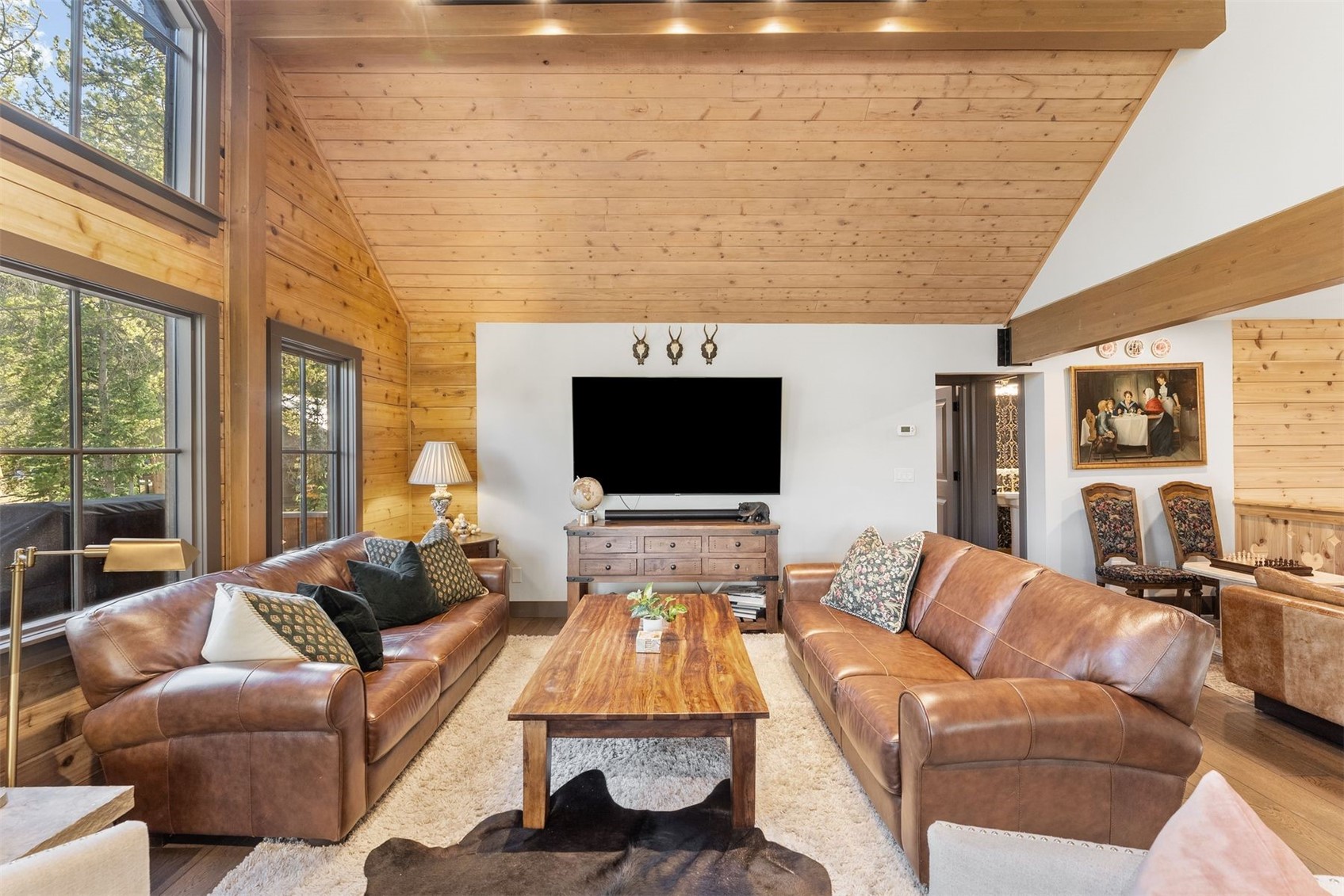 Living room with a vaulted wood ceiling, leather sofas, and tall windows overlooking trees.