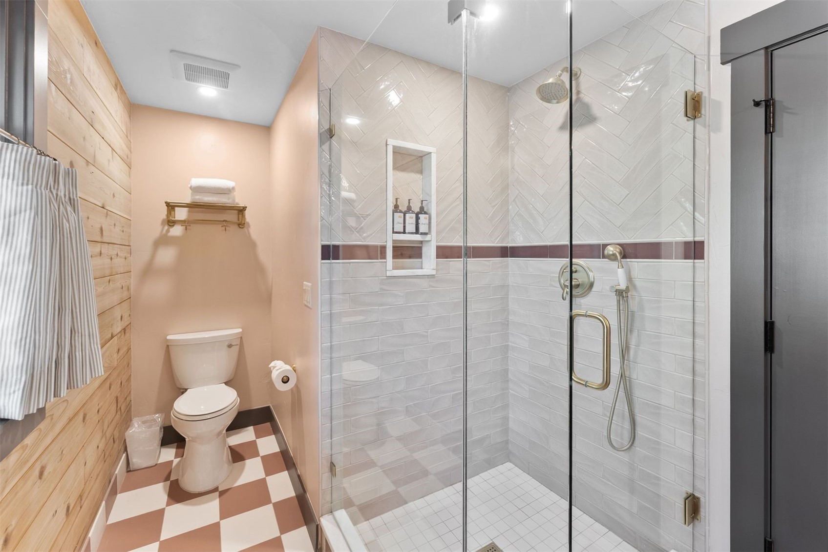 Bathroom with a glass walk-in shower, brass fixtures, wood-paneled wall, and pink-and-white checkerboard floor.
