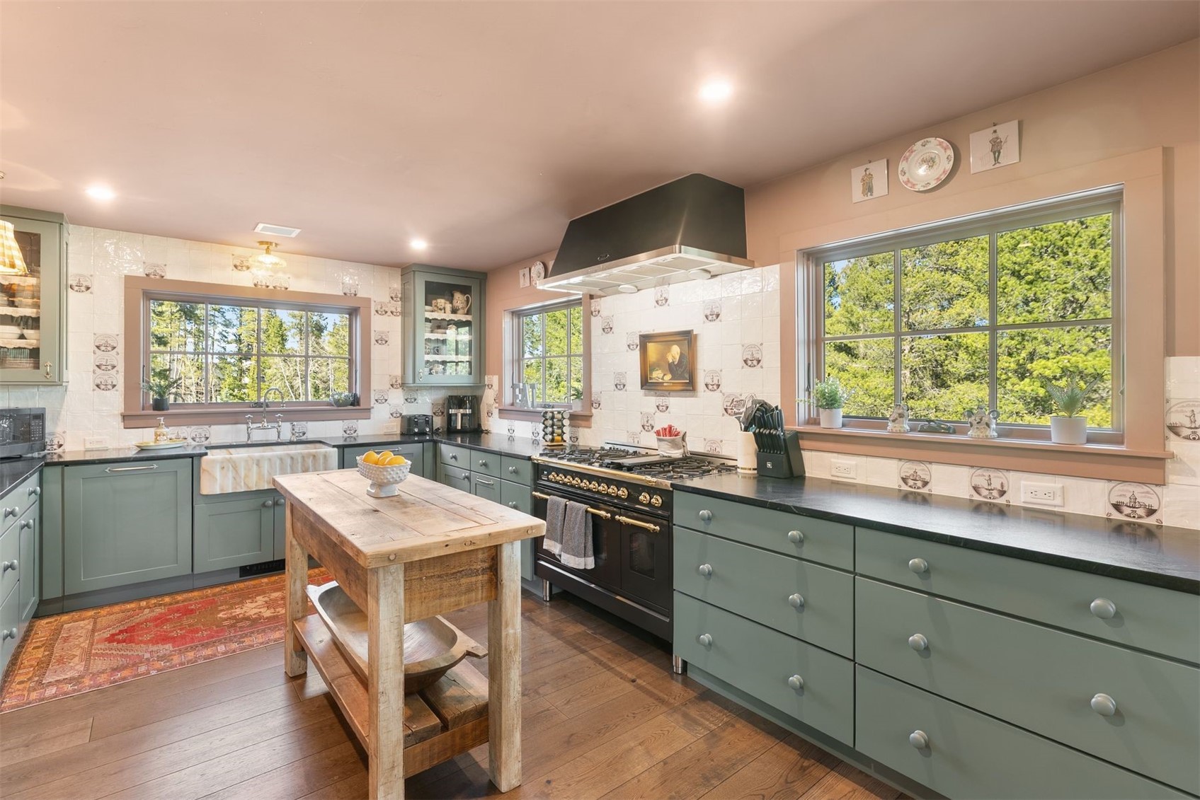 Kitchen with sage-green cabinets, a black range, rustic wood island, and large windows overlooking trees.