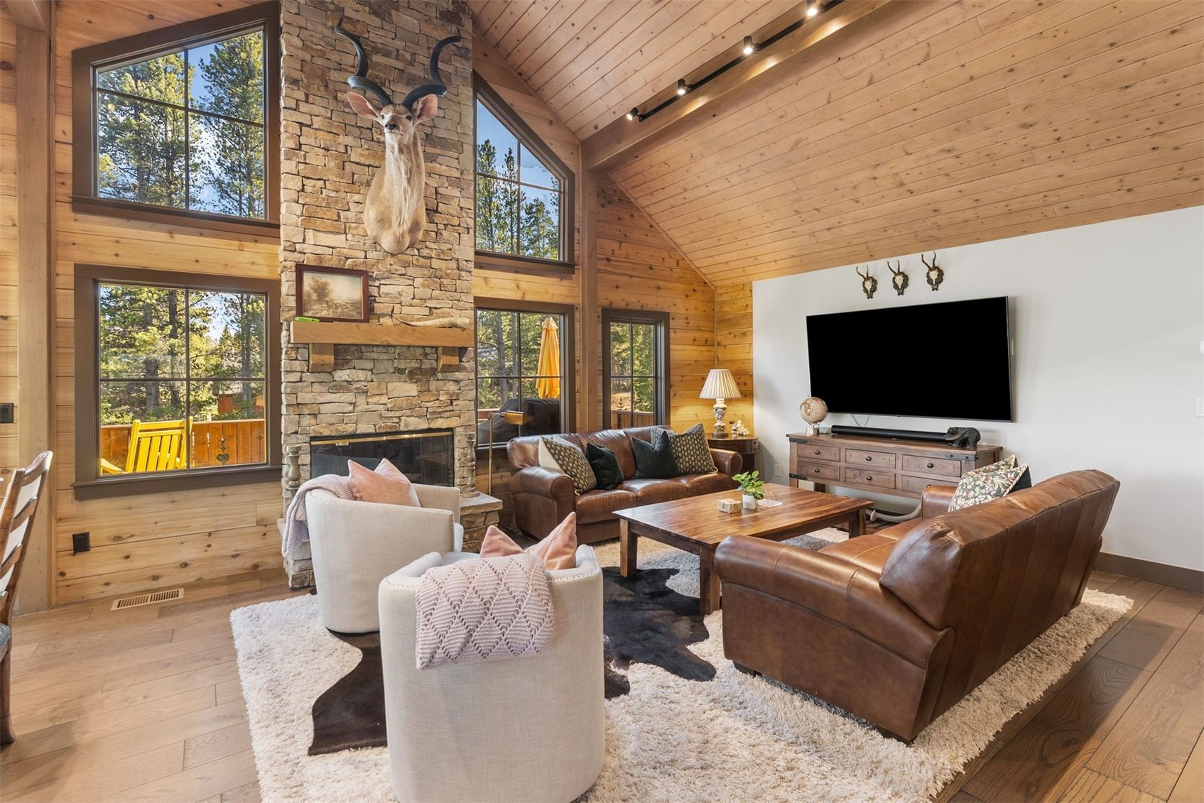 Living room with a vaulted wood ceiling, stone fireplace, leather sofas, and large windows overlooking trees.