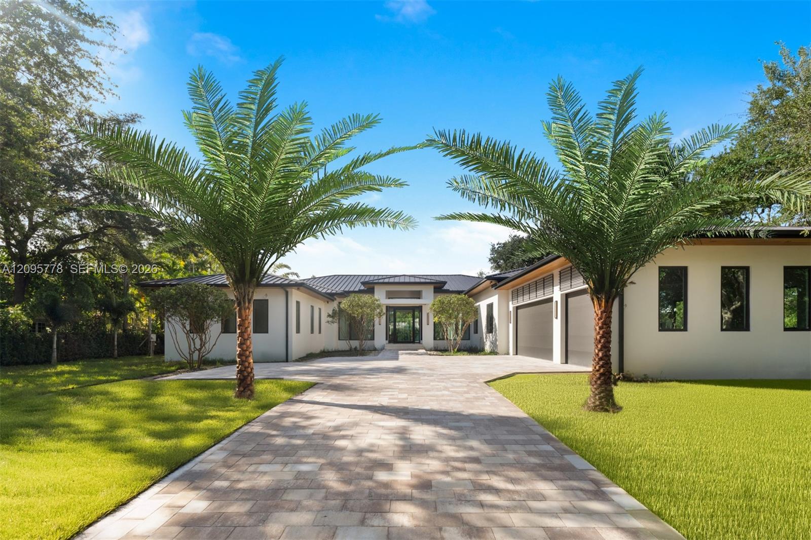 Modern white house with a dark metal roof, framed by palm trees beside a paved driveway.