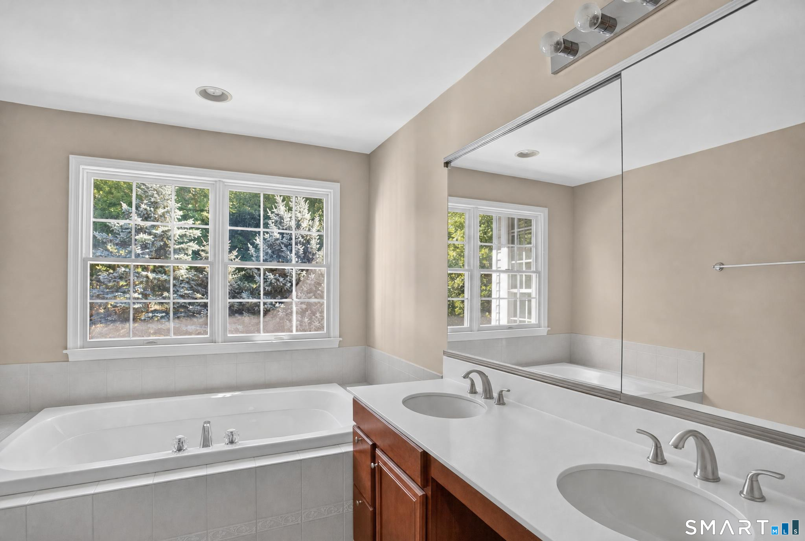 Bathroom with a soaking tub, double vanity, large mirror, and windows overlooking trees.
