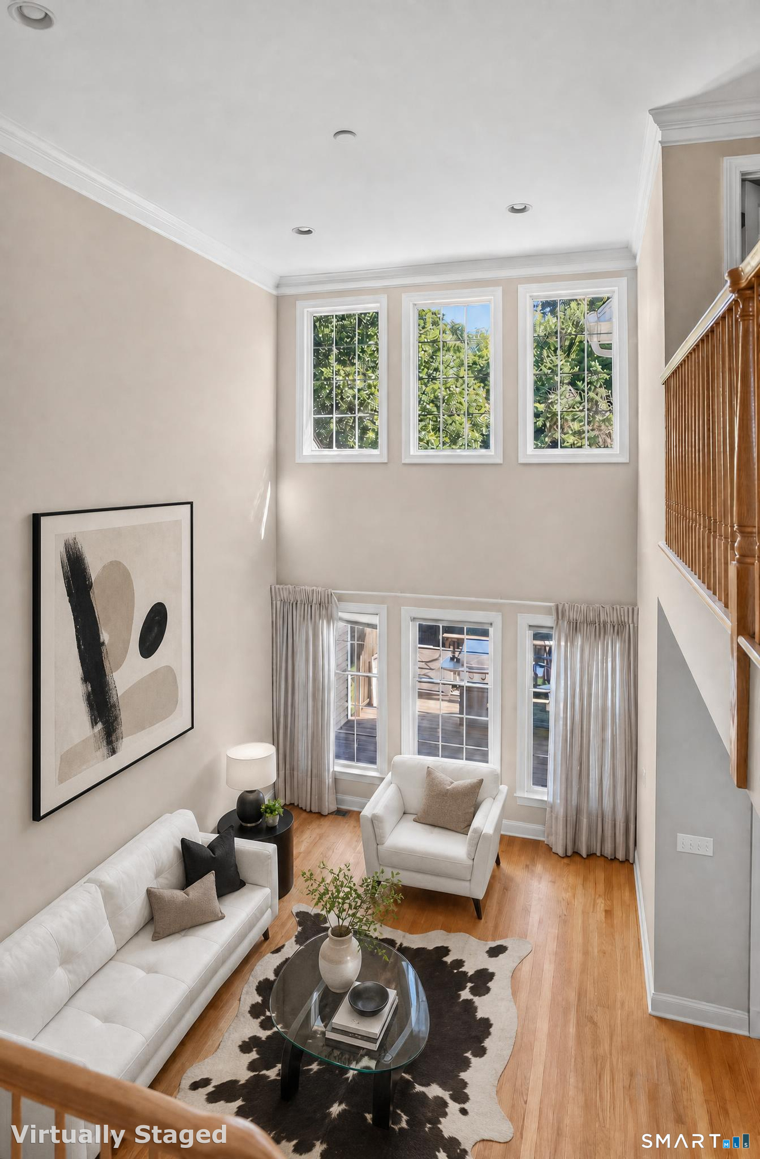 Double-height living room with white seating, tall windows, hardwood floors, and a wooden staircase.