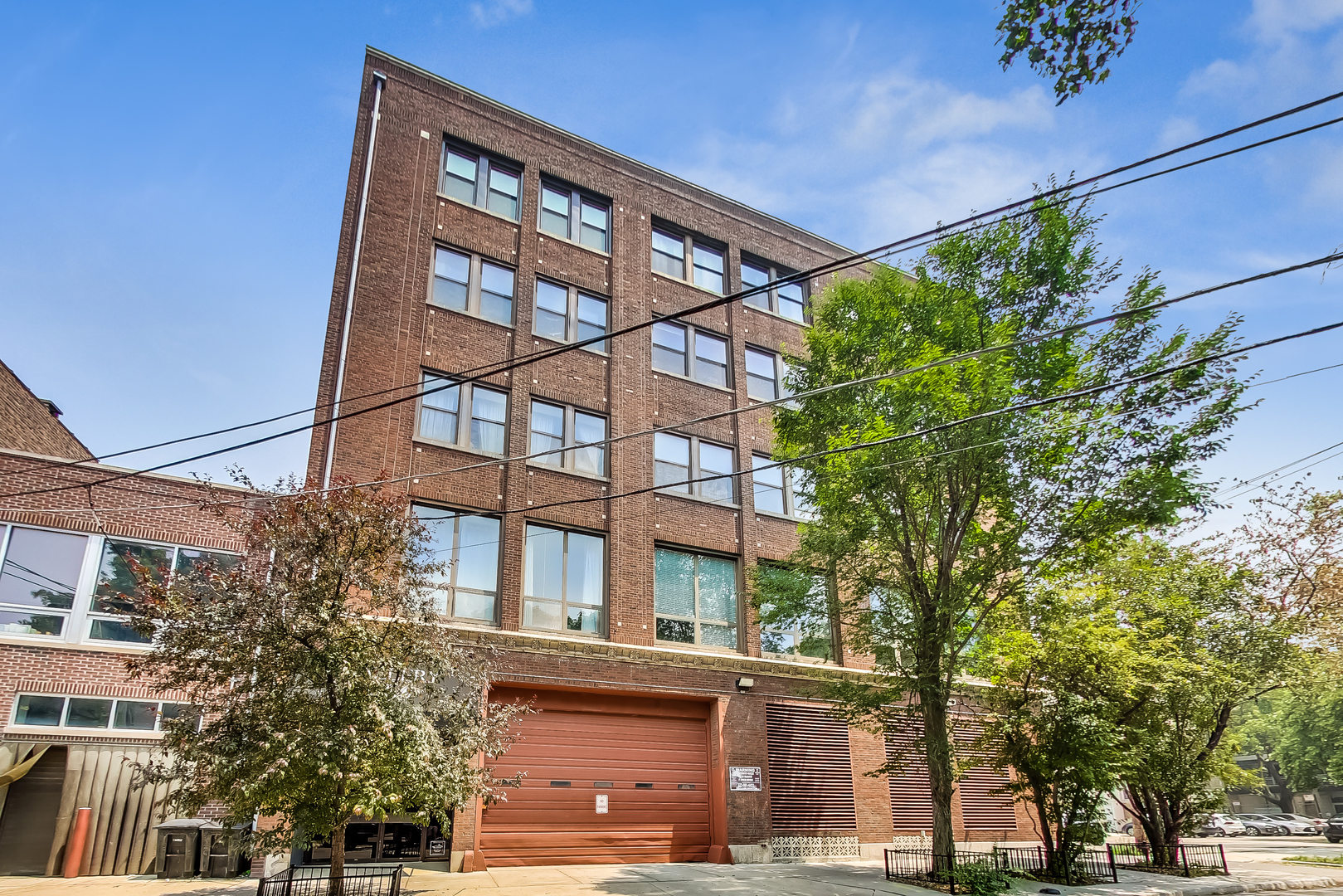 Multi-story brick residential building with rows of windows, a ground-level garage, and leafy street trees.