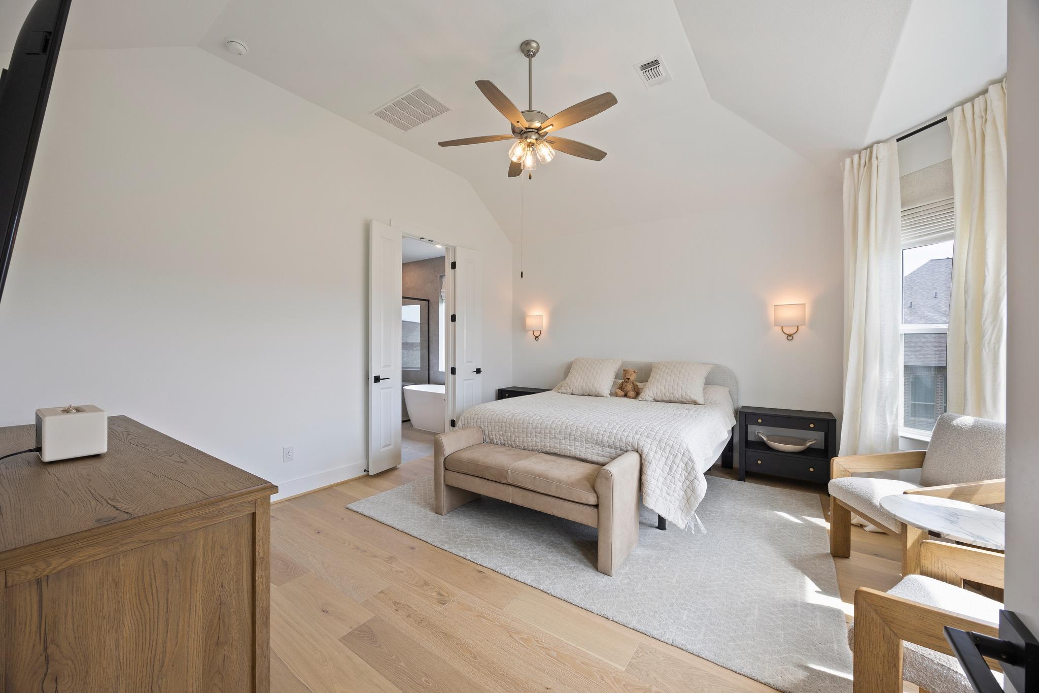 Furnished bedroom with vaulted ceiling, wood floors, ceiling fan, and a doorway to the bathroom.