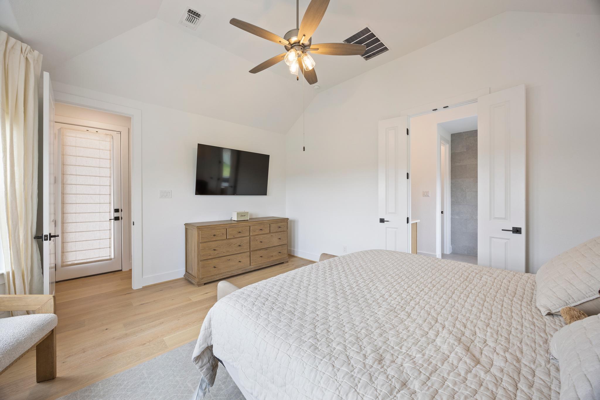 Bedroom with a white quilted bed, wooden dresser, wall-mounted TV, and ceiling fan.