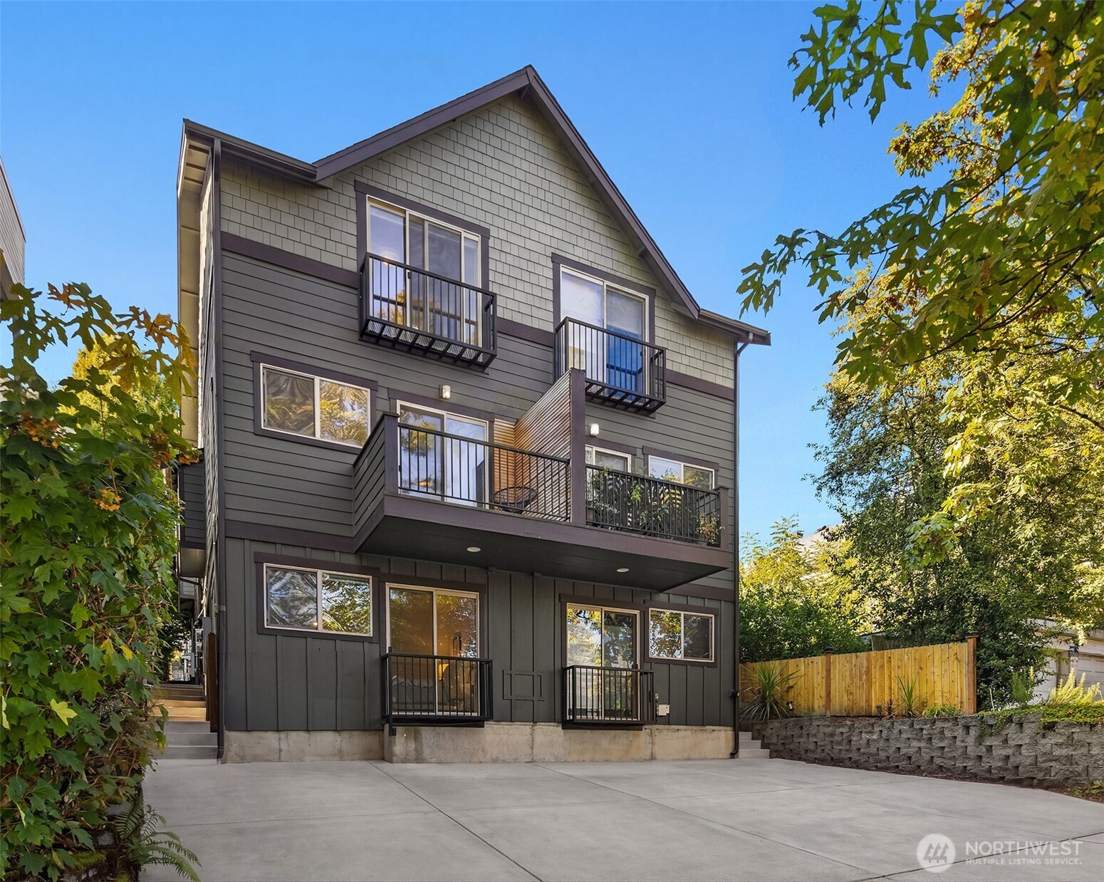 Multi-story gray home with several balconies, a wide concrete driveway, and autumn trees.