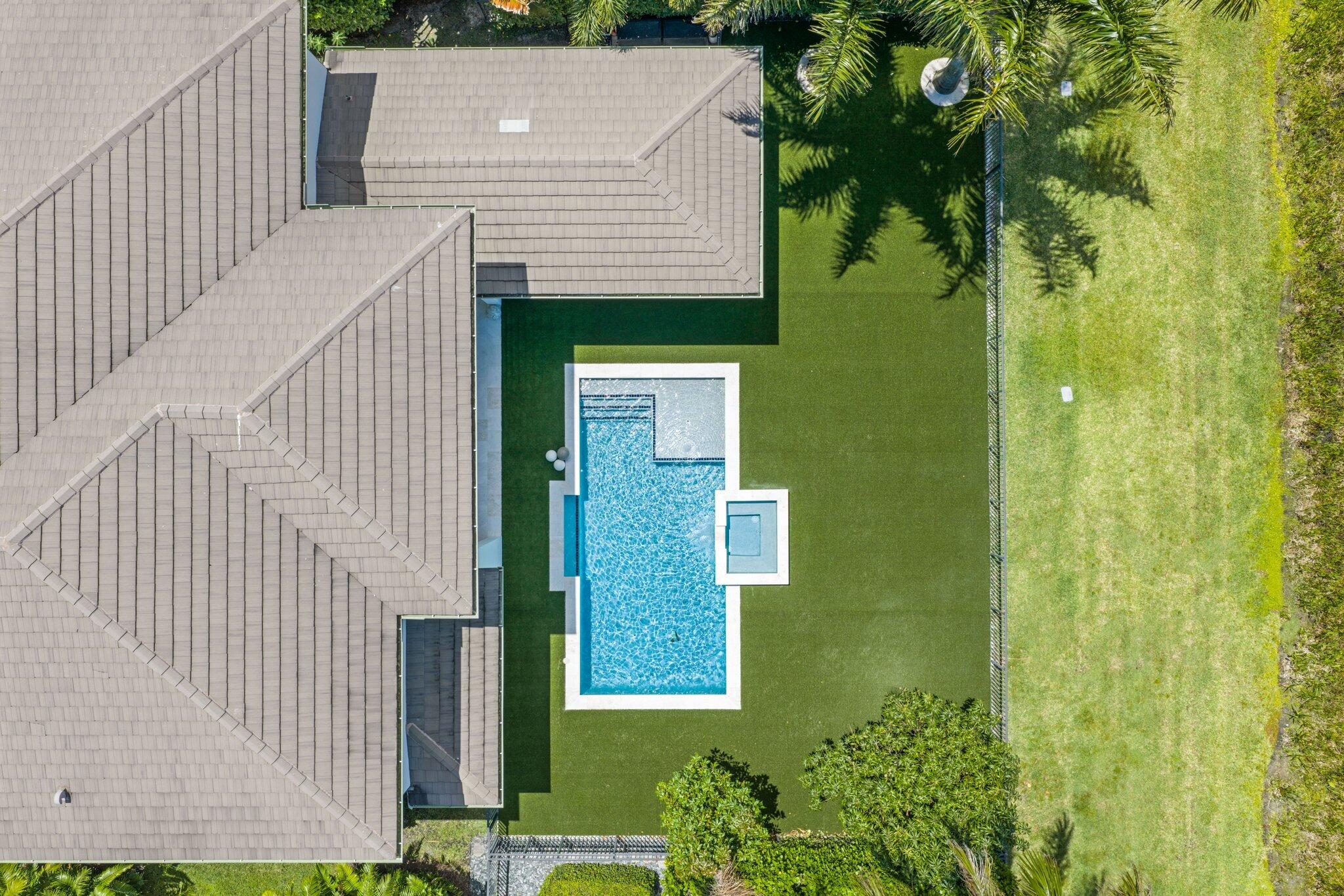 Aerial view of a backyard pool and spa beside a gray tiled roof, with a broad green lawn and palm trees.