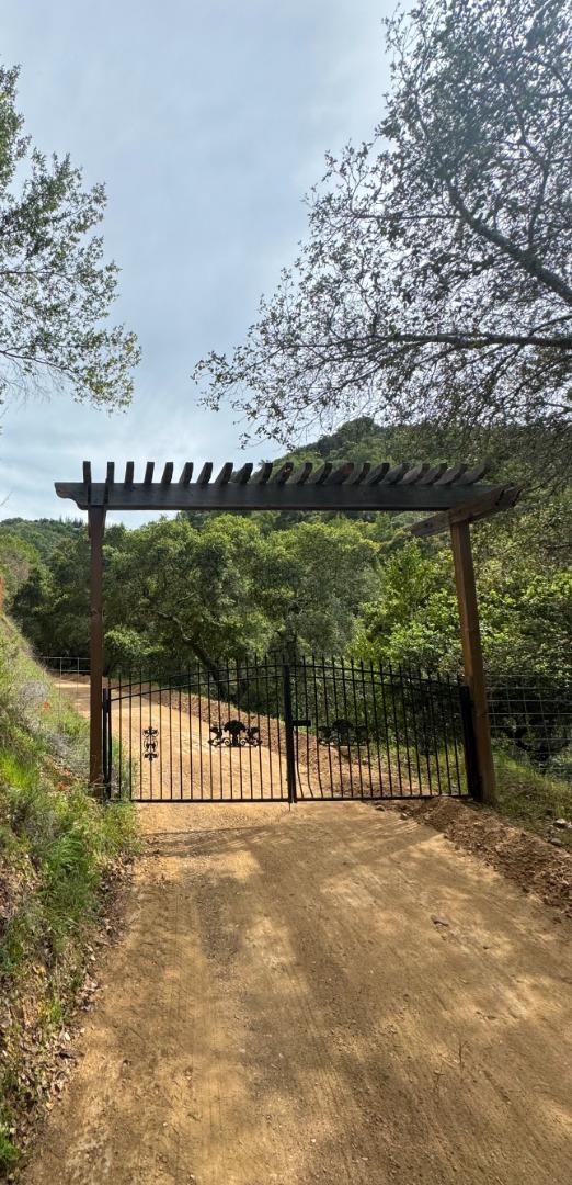 Gated dirt entrance to a wooded property beneath a wooden pergola.