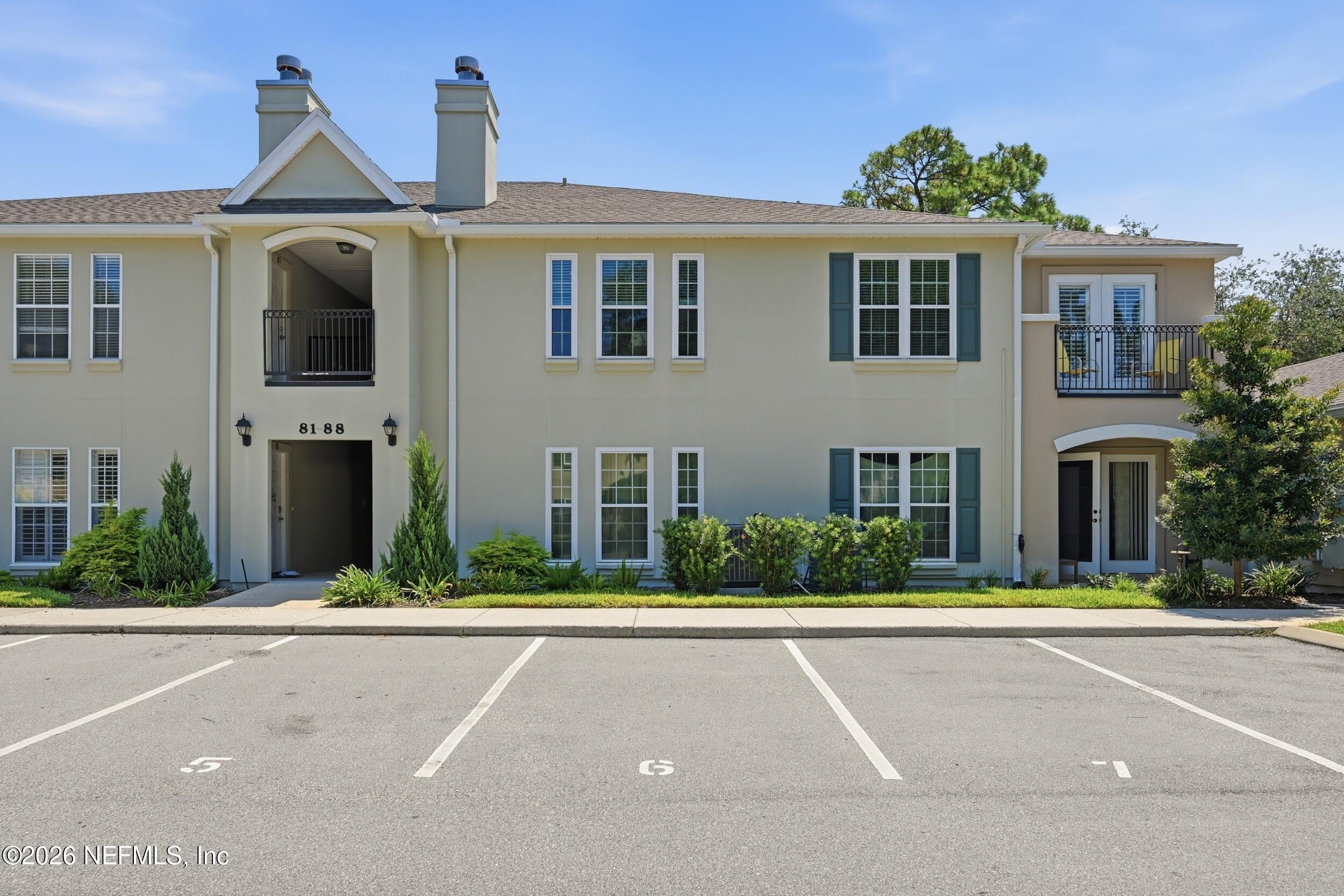 Two-story beige residential building with balconies, blue-green shutters, and parking spaces in front.
