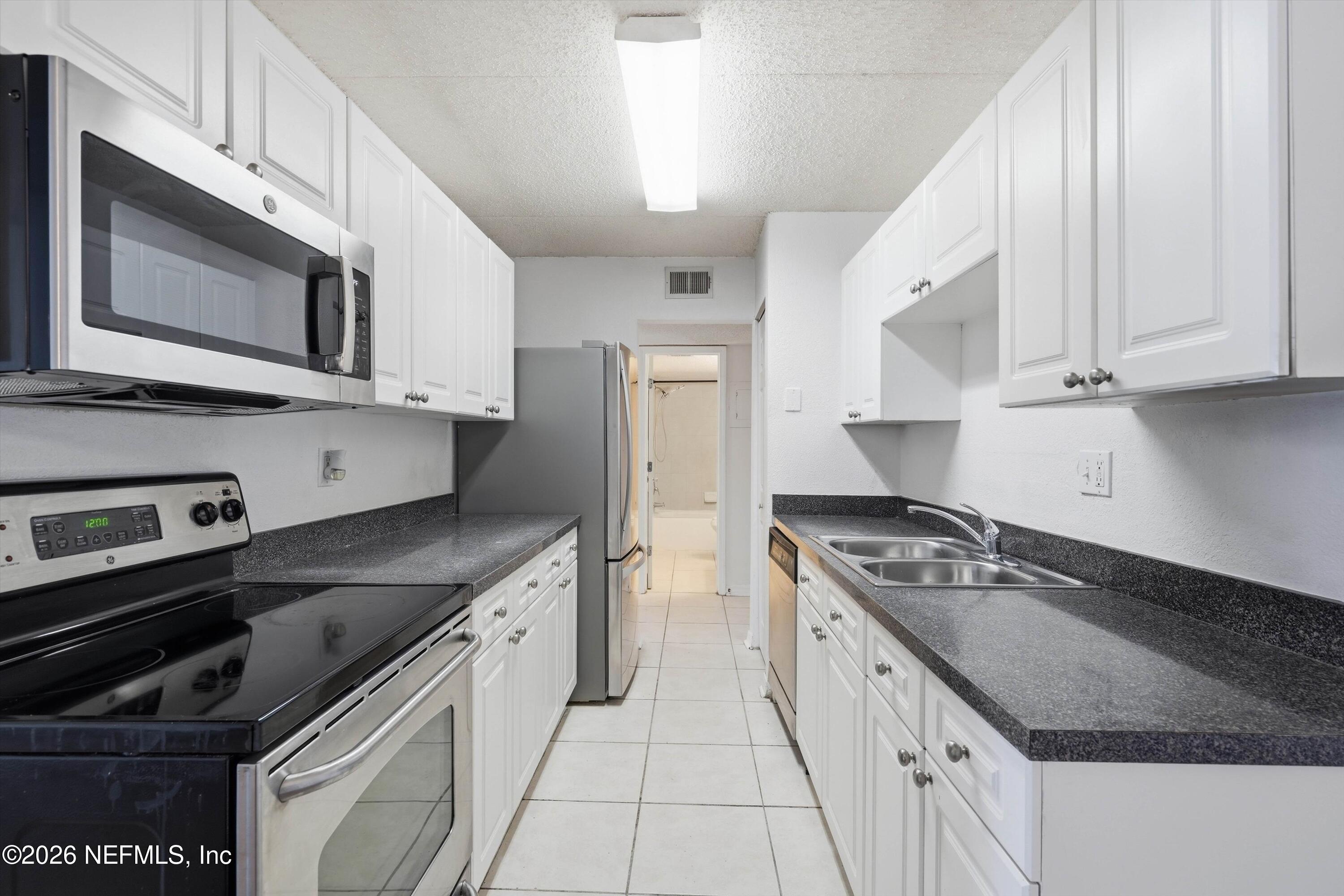 Galley kitchen with white cabinets, dark countertops, stainless appliances, and a double-basin sink.