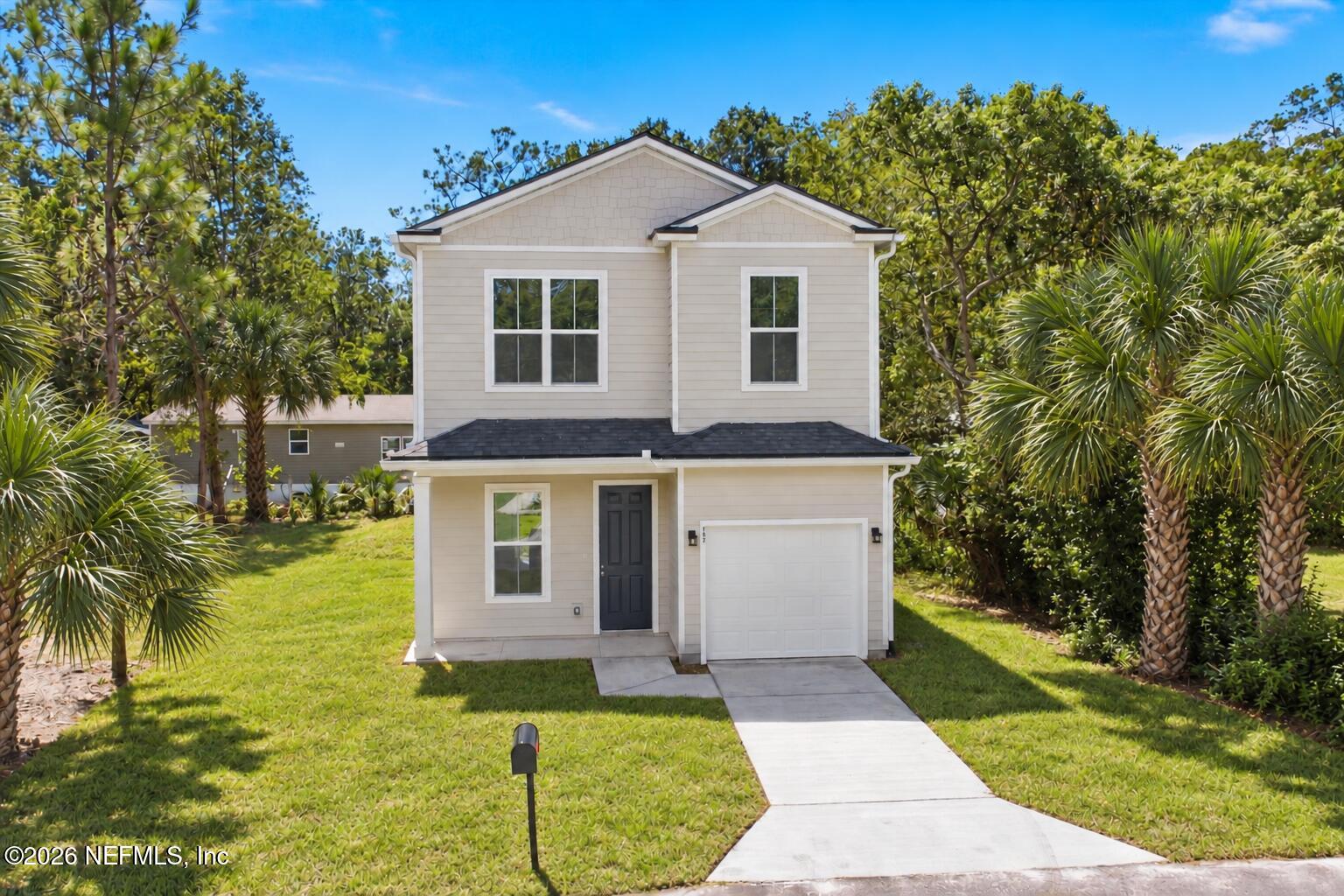 Two-story light-colored house with an attached garage, driveway, lawn, and tropical landscaping.