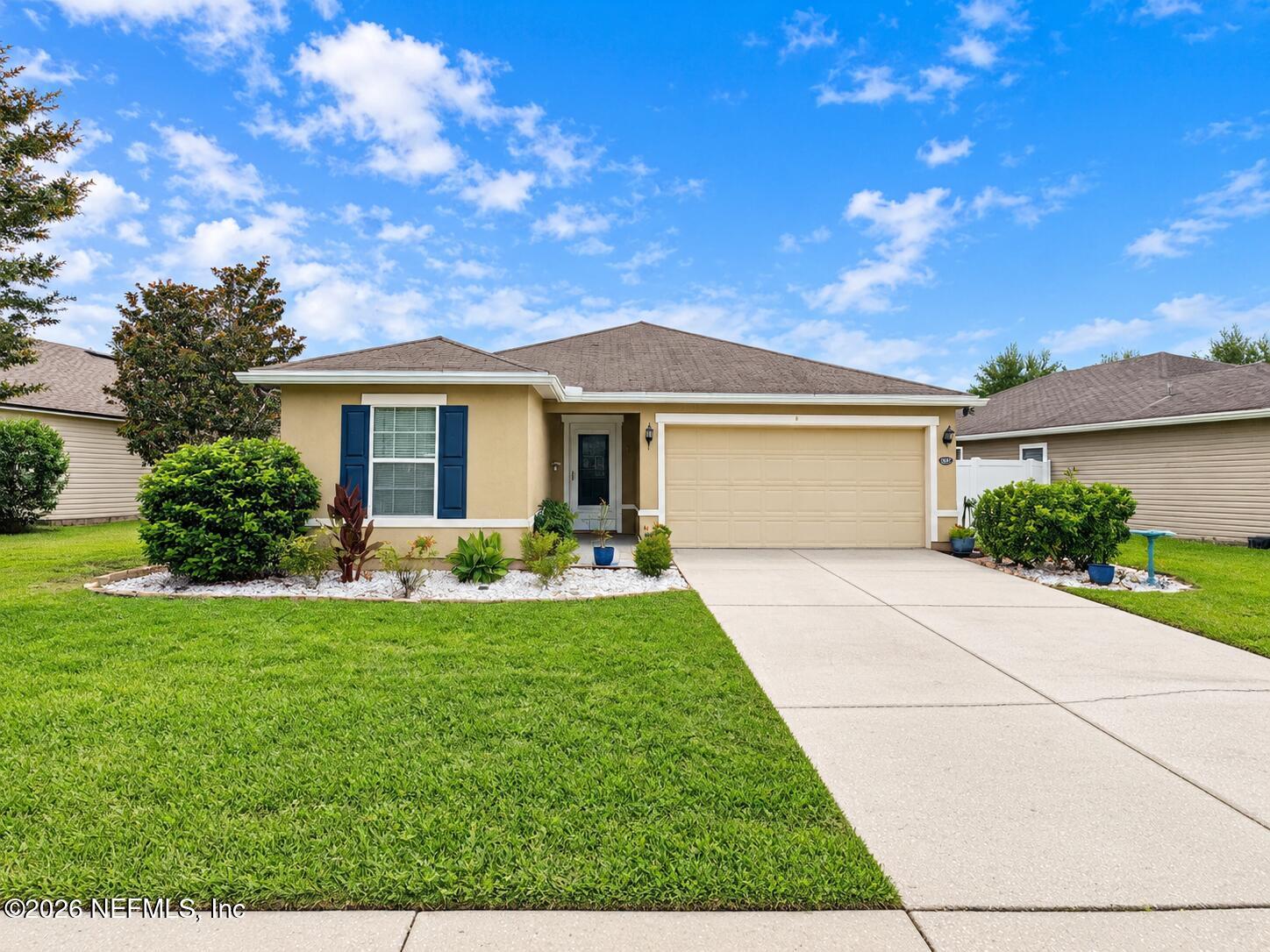 Single-story tan home with blue shutters, an attached garage, landscaped lawn, and concrete driveway.