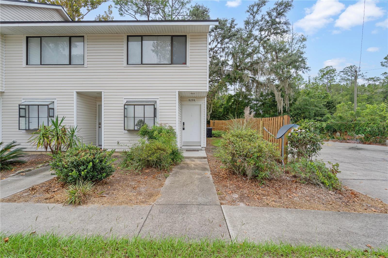 Two-story light-sided home with a covered entry, front walkway, shrubs, and wooden side fence.
