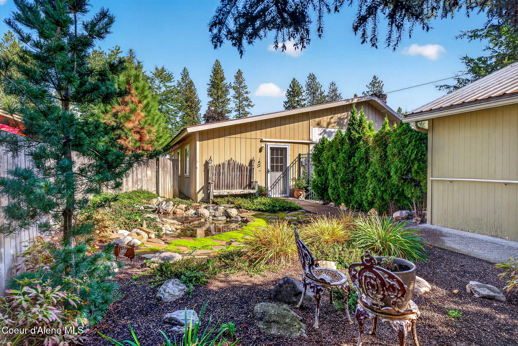 Landscaped backyard with a rock-lined water feature and ornate patio chairs beside a tan outbuilding.