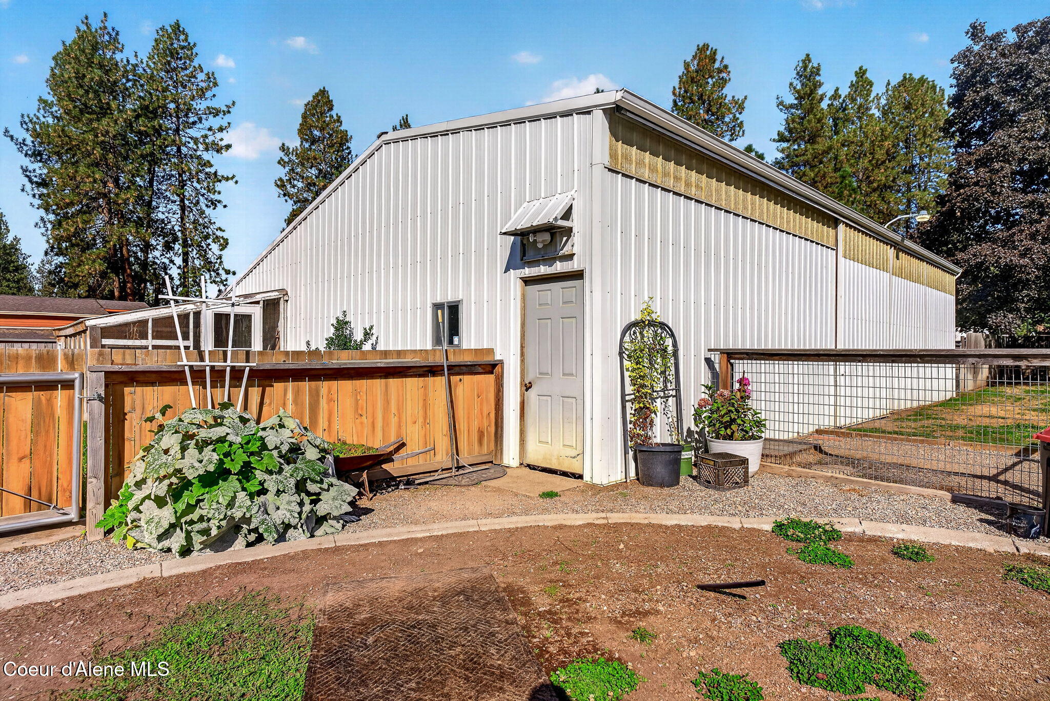 White metal outbuilding with a gray door, fenced garden beds, and gravel yard.