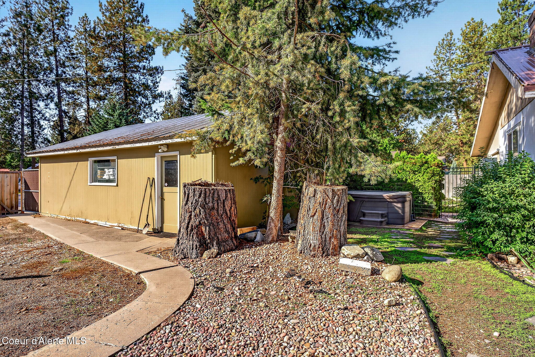Backyard with a tan outbuilding, hot tub, mature evergreens, and a curved concrete walkway.