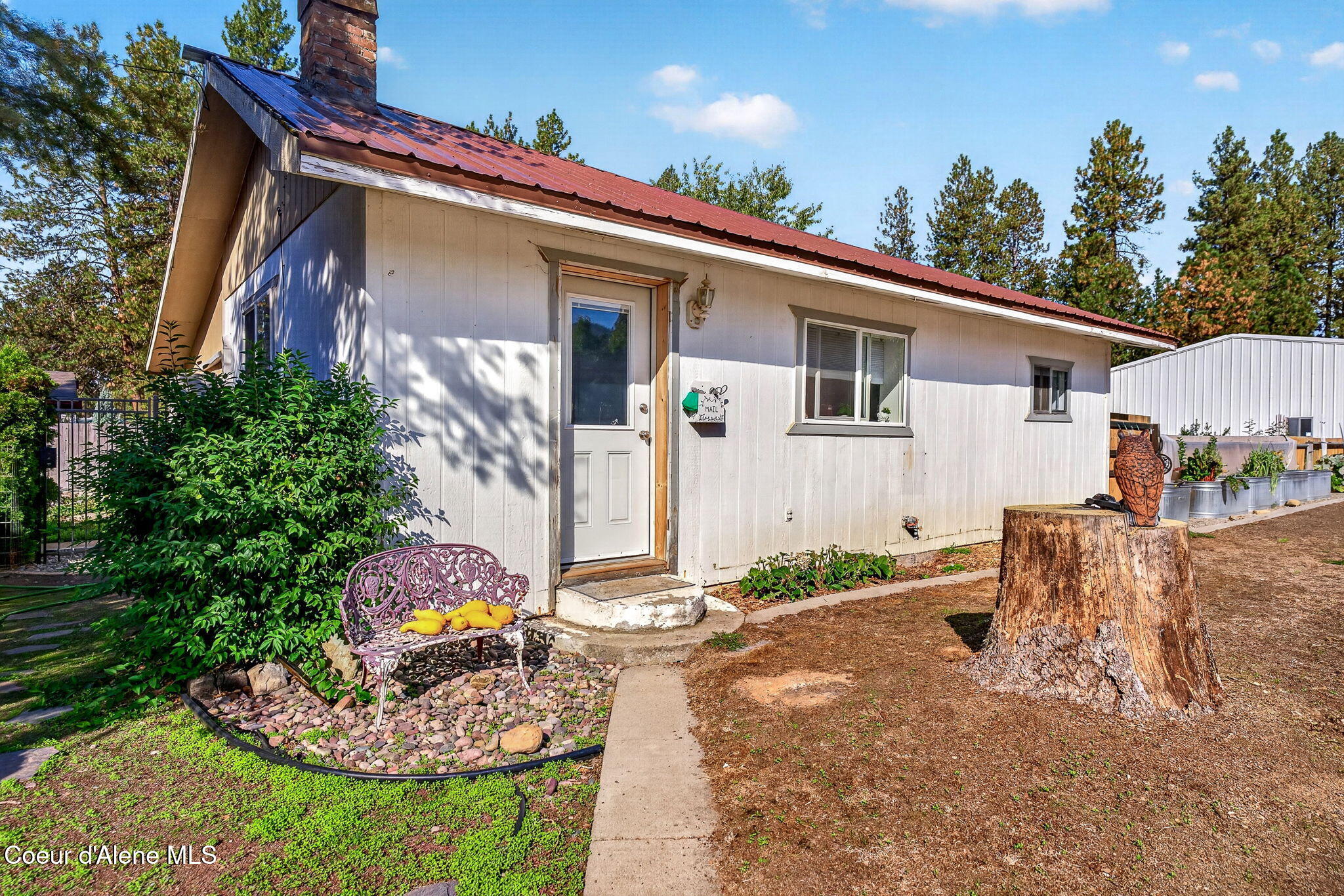 White-sided home with a red metal roof, brick chimney, garden bench, and large tree stump in the yard.