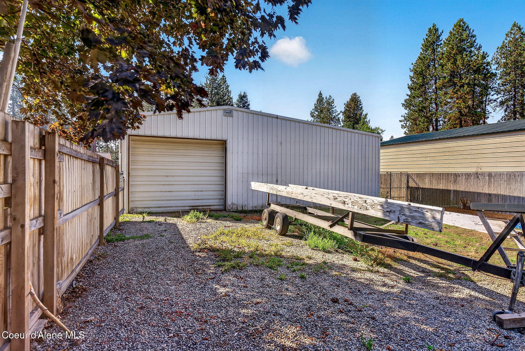 Detached metal garage with roll-up door in a fenced gravel yard beside a boat trailer.