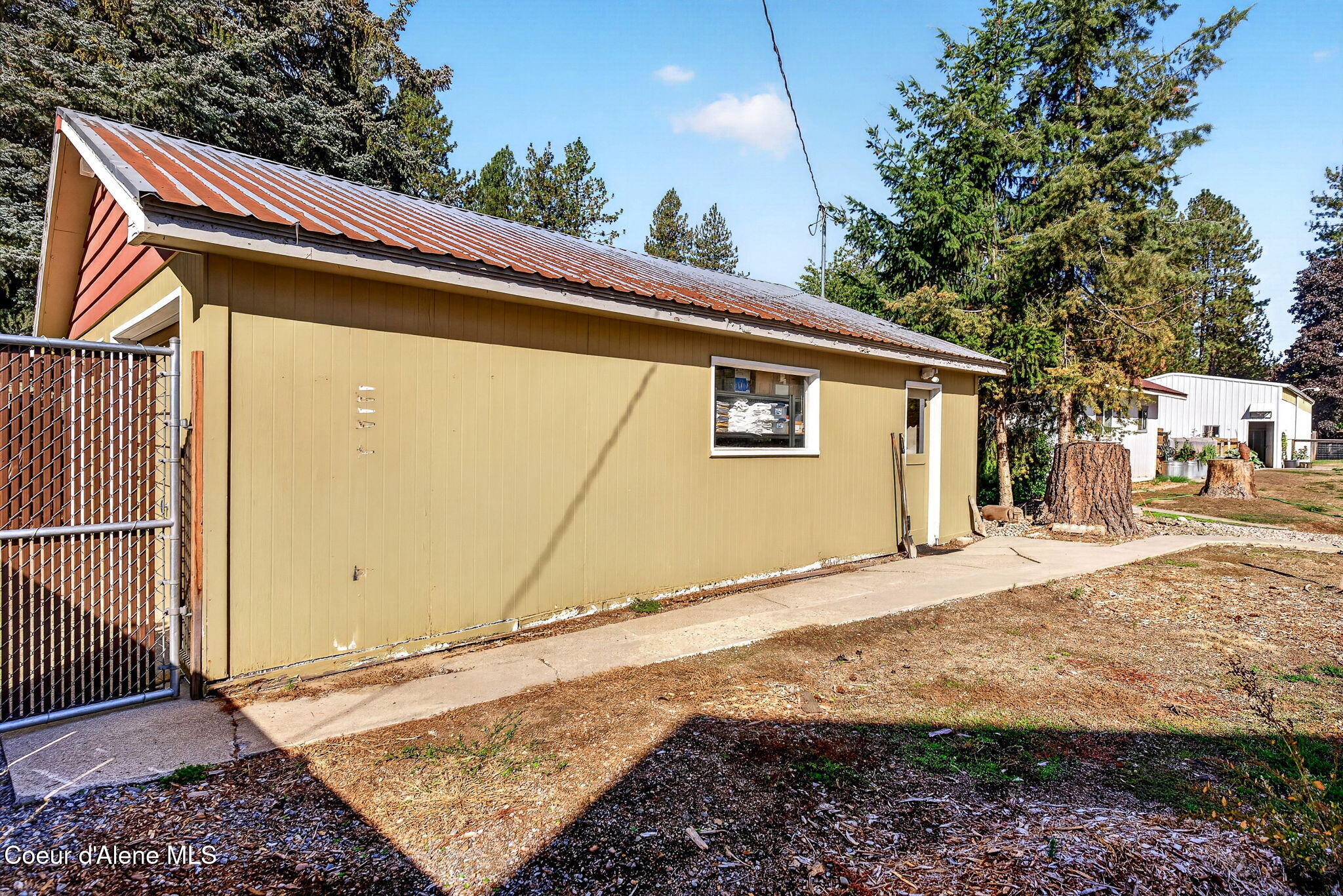 Tan outbuilding with a weathered metal roof beside a chain-link gate and concrete walkway.