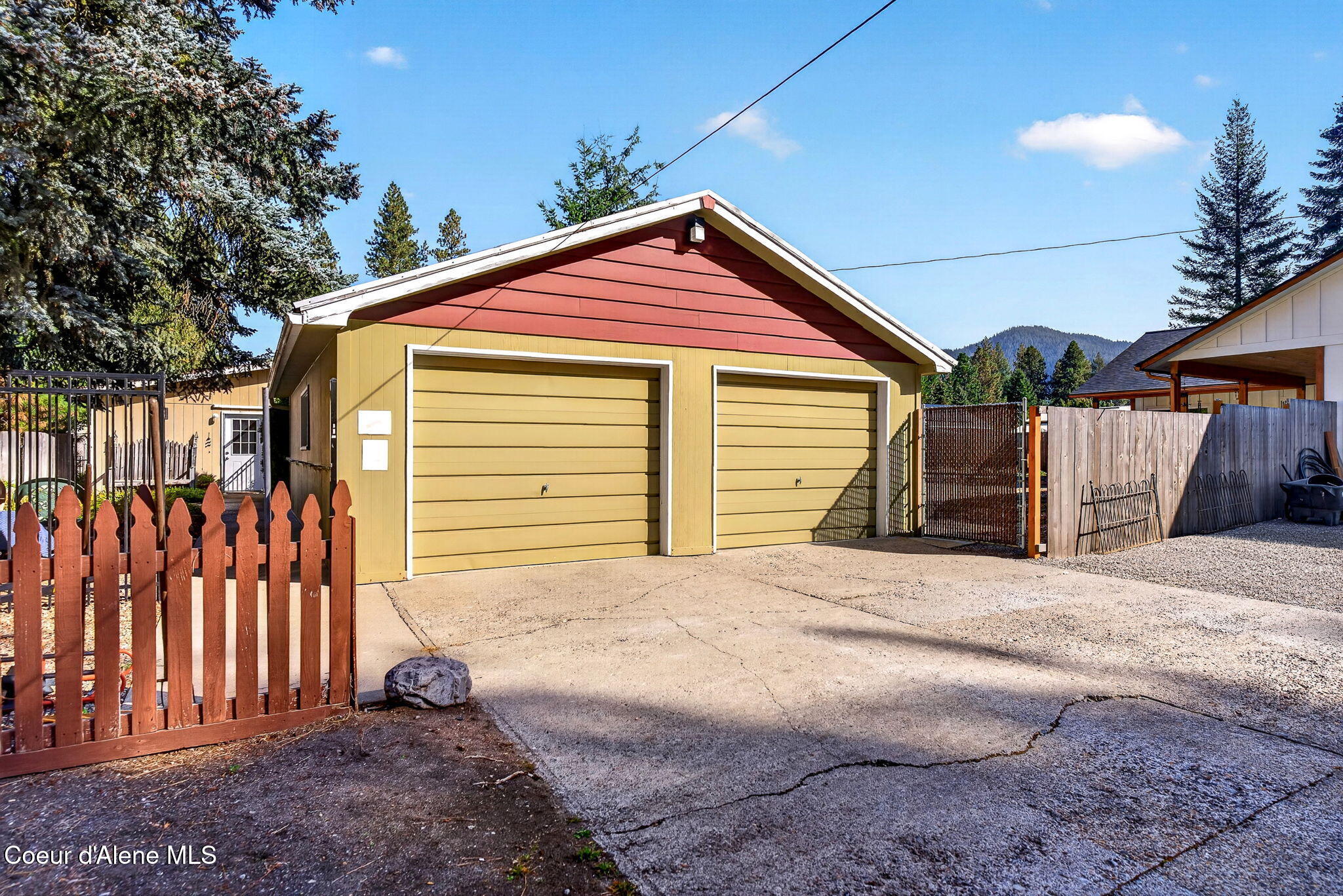 Yellow two-car garage with a red gable, fenced driveway, and wooded mountain in the background.
